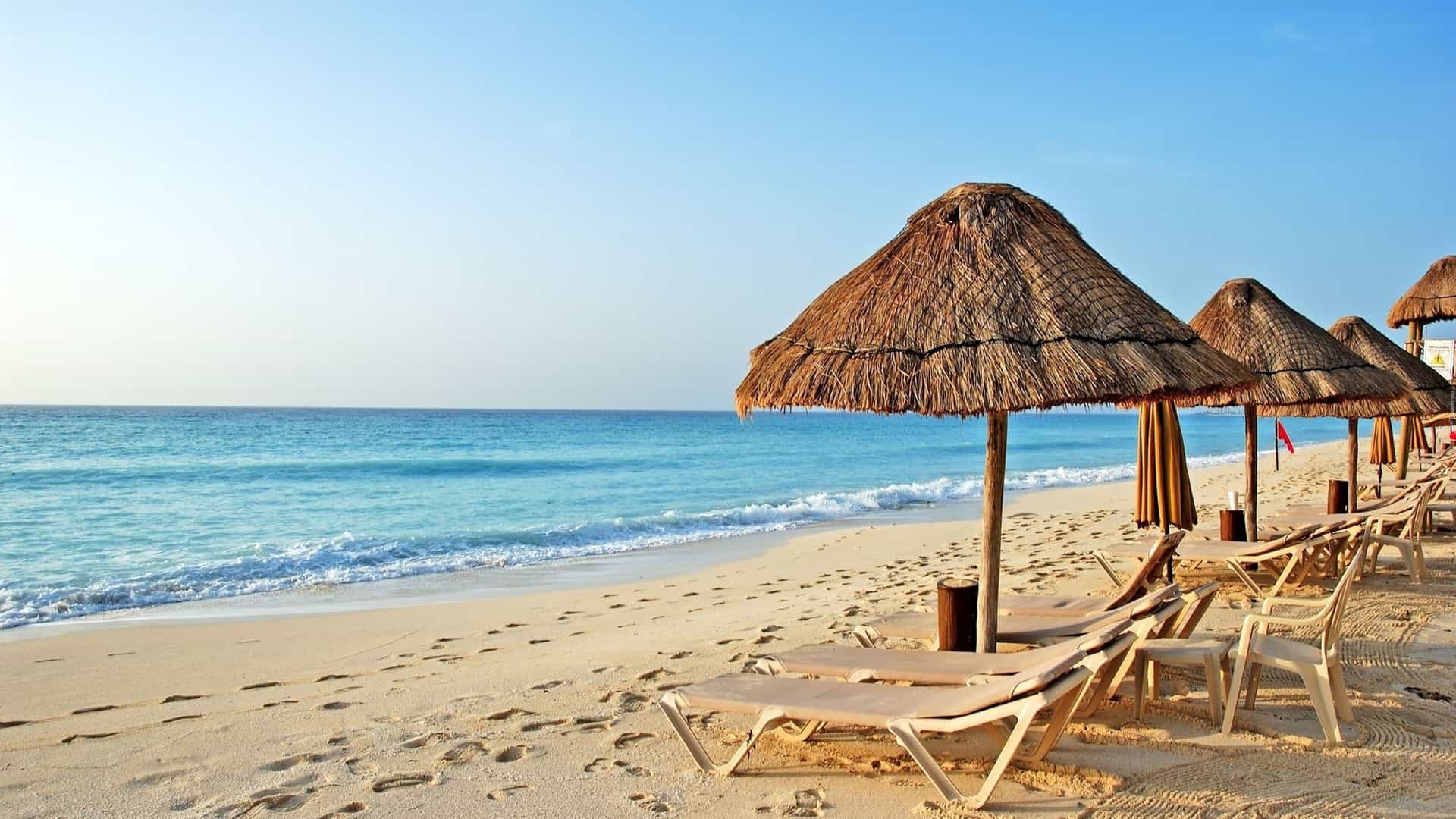 A peaceful beach scene in the Caribbean with lounge chairs and thatched umbrellas lined up along the white sand. The chairs face the calm, turquoise water of the ocean, which gently laps the shore in small, white waves. The sand is scattered with footprints, and the sky is a clear, light blue. The image captures a classic and tranquil view of a tropical beach, a key feature of the Caribbean region.