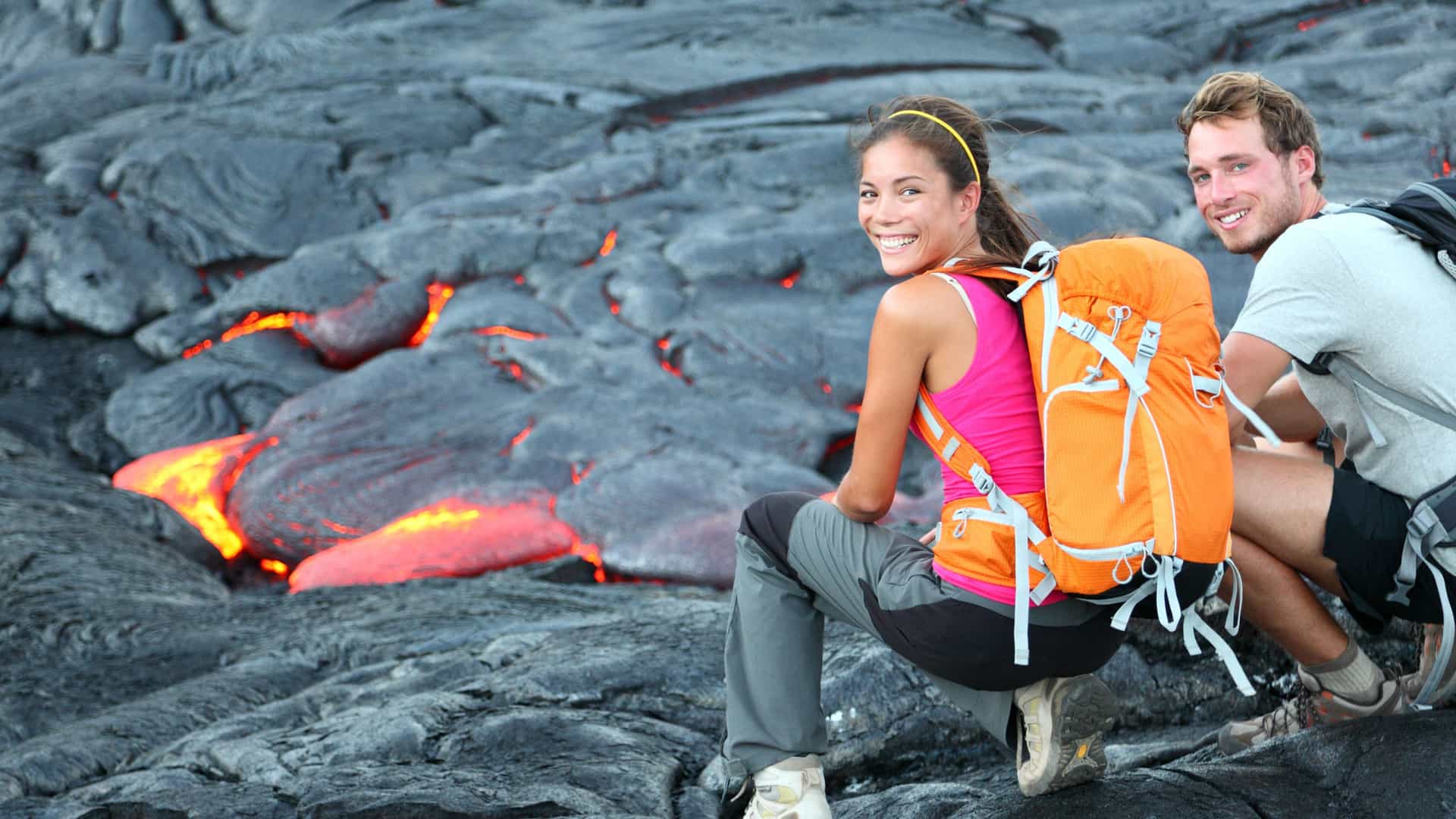 A smiling couple, a man and a woman, are sitting on a bed of cooled, black lava rock during a volcano tour in Hawaii. In the background, bright orange molten lava glows from cracks and crevices in the rock. The woman, wearing a pink shirt and a bright orange backpack, looks back at the camera with a happy expression. The man is also smiling. The image captures the unique experience of hiking on an active volcanic landscape in the Hawaii region.