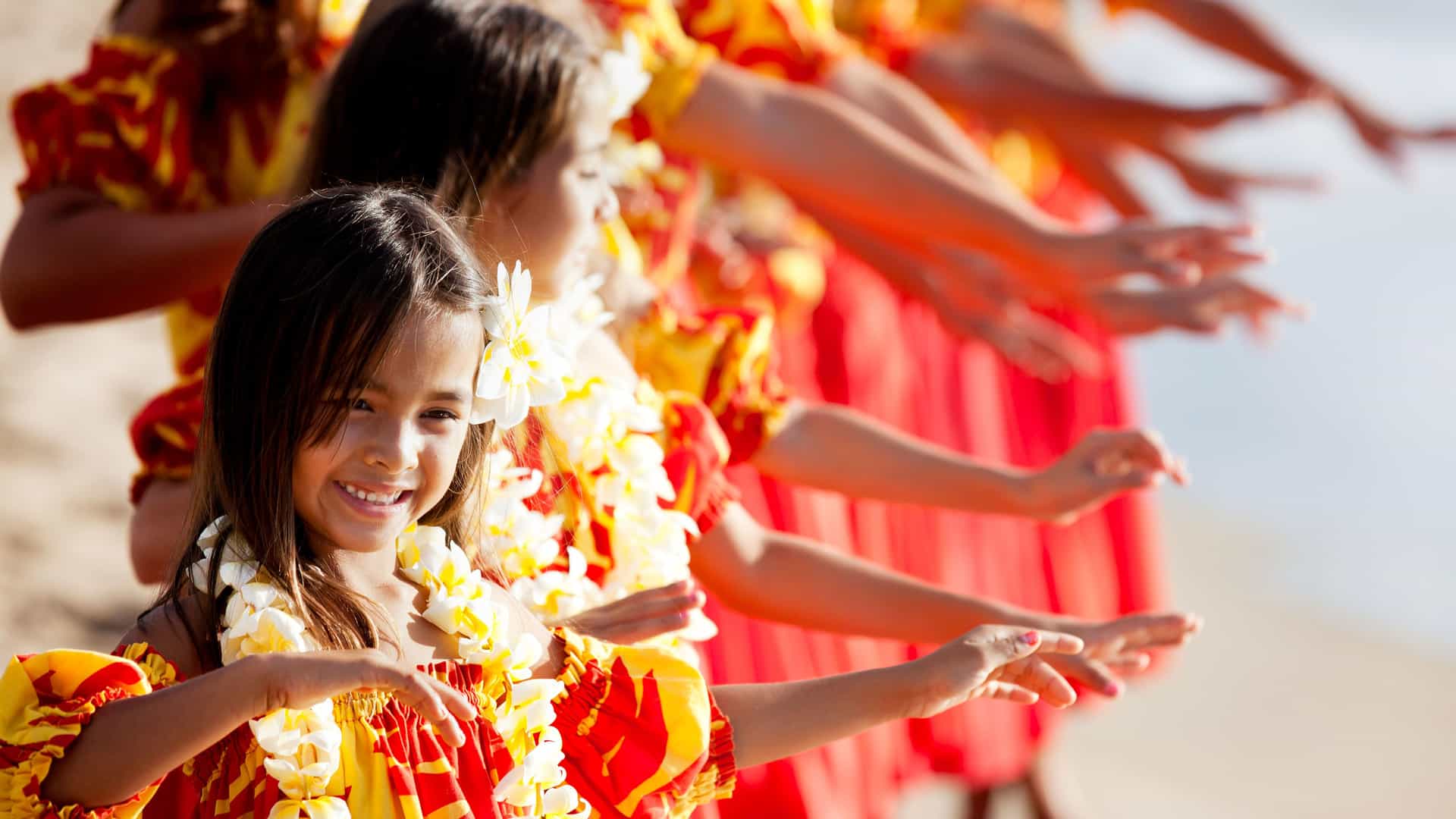 A group of smiling Hawaiian hula dancers, including a young girl in the foreground, perform on a beach. They are wearing traditional red and yellow floral-patterned dresses and white flower leis around their necks. They are positioned in a line, with their arms extended in a graceful hula movement. The background is a soft blur of sand and ocean, capturing a classic cultural experience of the Hawaii region.