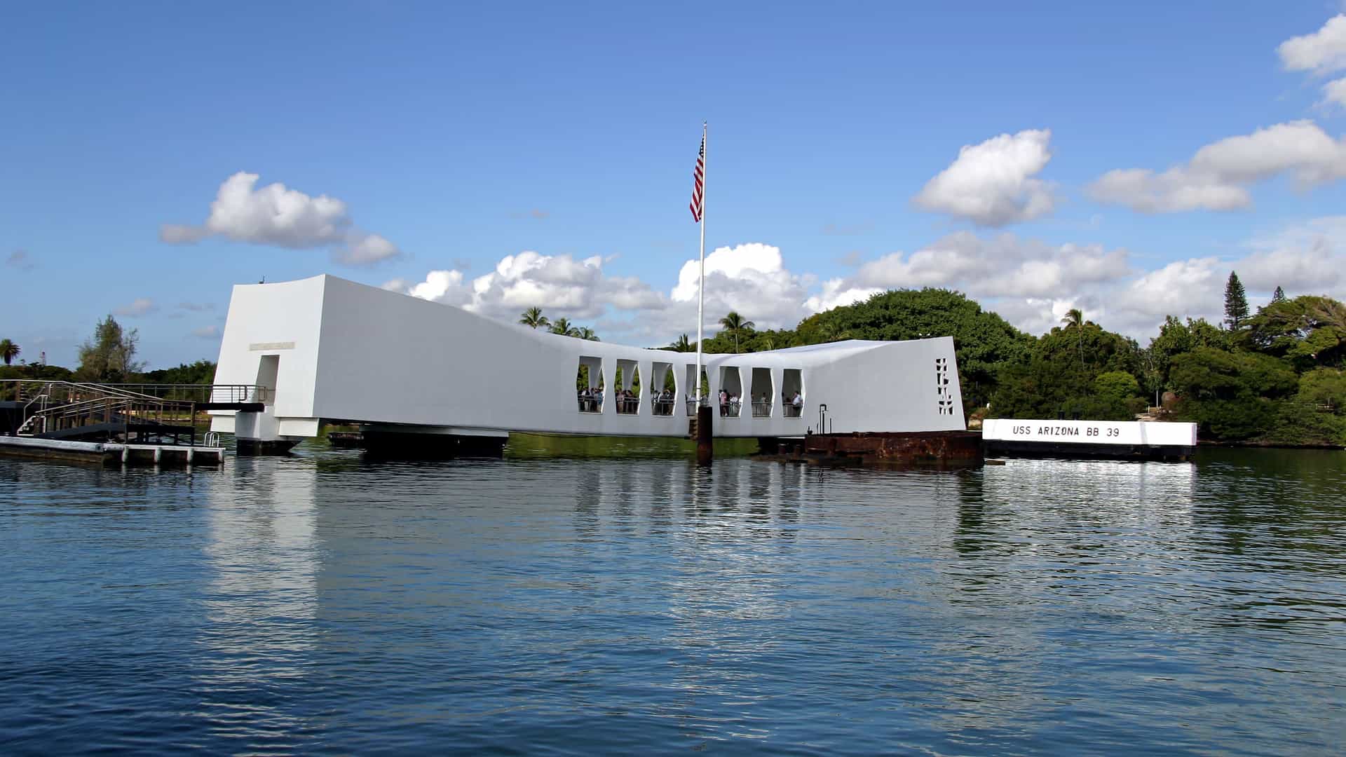 A view of the USS Arizona Memorial at Pearl Harbor in Hawaii. The memorial is a stark, white, floating structure with an open, bridge-like design, built directly over the sunken battleship USS Arizona. A United States flag flies from a mast positioned at the center of the memorial. The water is calm and dark, with the memorial and the sky reflected in its surface. In the background, lush green trees line the shore under a blue sky with scattered white clouds, capturing the solemn and historically significant location of the Hawaii region.