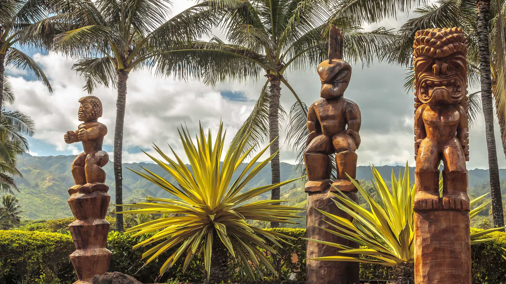 Three traditional Polynesian tiki statues, carved from wood, stand in a lush Hawaiian landscape on Oahu. The carvings depict stylized human-like figures with intricate facial expressions. Palm trees and large, spiky yellow and green plants surround the statues. In the background, green, mist-covered mountains are visible under a cloudy sky, showcasing the rich cultural art and natural beauty of the Hawaii region.