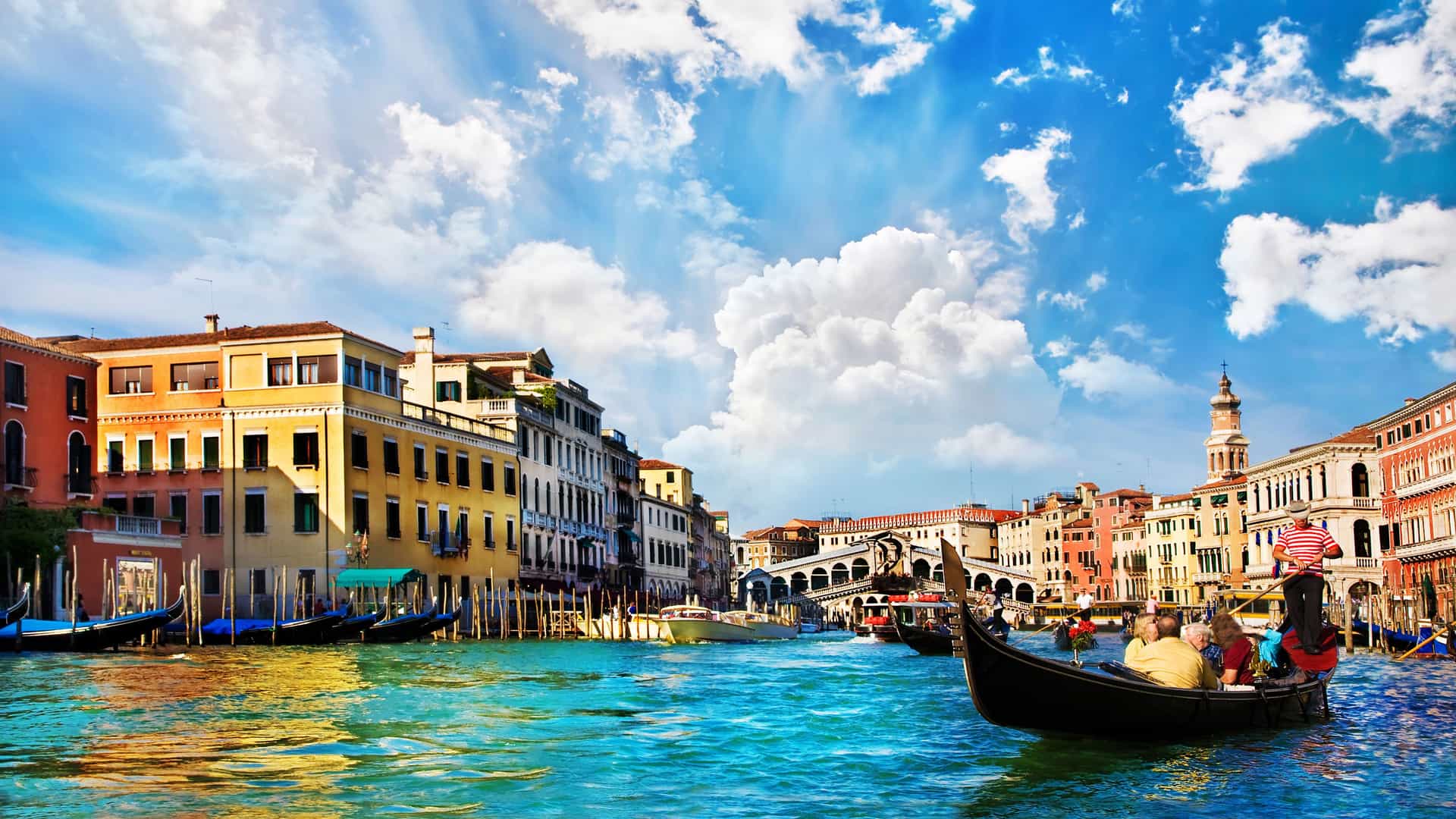 A gondola carrying tourists glides along the turquoise-blue waters of the Grand Canal in Venice, Italy. The iconic Rialto Bridge is visible in the background, connecting two rows of historic, colorful buildings. Other gondolas are moored along the canal banks. The sky is bright blue with large, white, fluffy clouds, creating a classic, picturesque scene of this Mediterranean city.