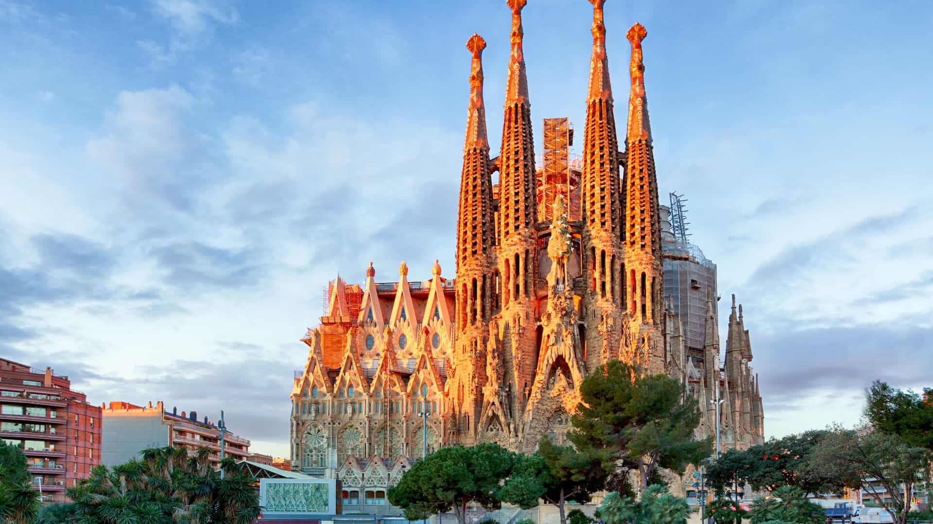 The iconic Sagrada Familia cathedral in Barcelona, Spain, bathed in the warm, golden light of sunset. The intricate facade and towering spires of the Gaudi masterpiece are highlighted by the low sun. In the foreground, a tranquil pond reflects the sky and the surrounding trees, and modern buildings are visible on the left. The sky is a mix of deep blue and wispy clouds, creating a dramatic and beautiful scene of this famous Mediterranean landmark.