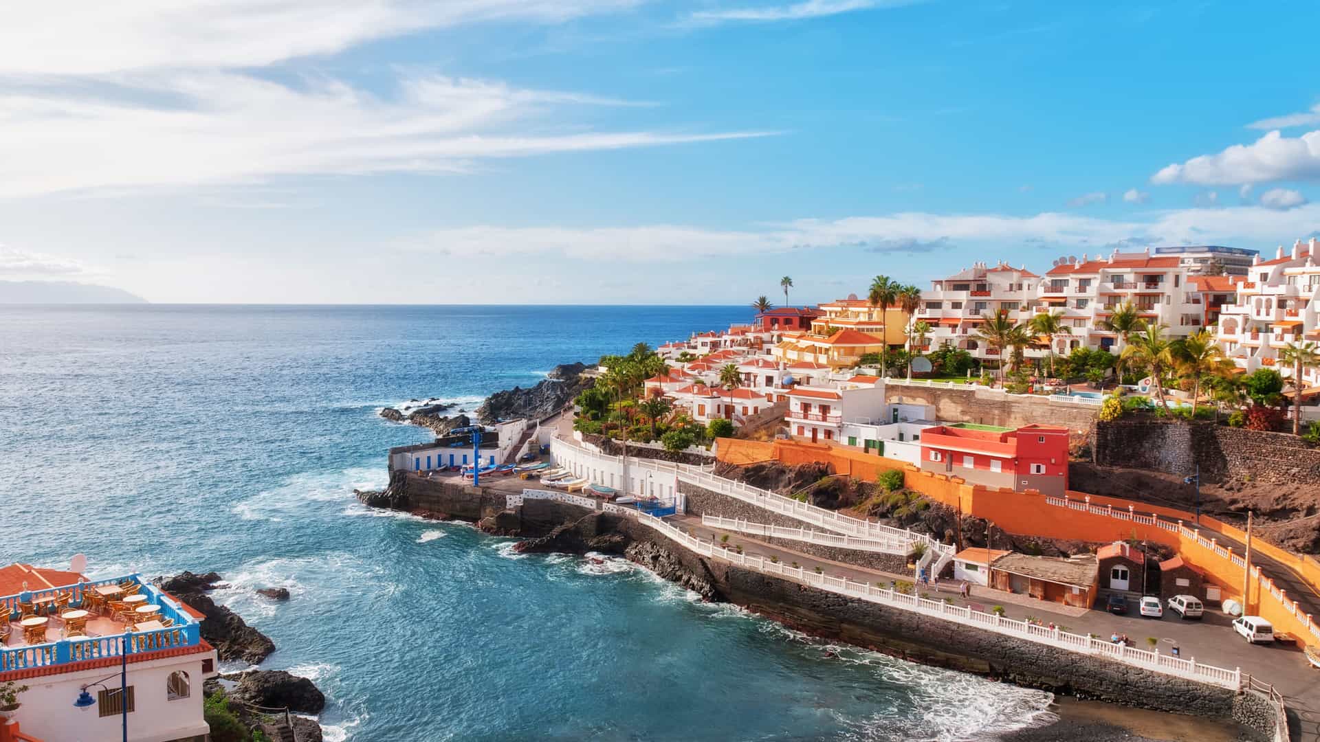 An elevated view of Puerto Santiago on the coast of Tenerife in the Canary Islands, Spain. A collection of white and orange buildings with red roofs are built into a cliffside overlooking the Atlantic Ocean. A paved walkway follows the rocky coastline, which leads to a small beach. The deep blue ocean has gentle waves, and the sky is bright blue with scattered white clouds. The image captures the beautiful coastal landscape of this Mediterranean region.