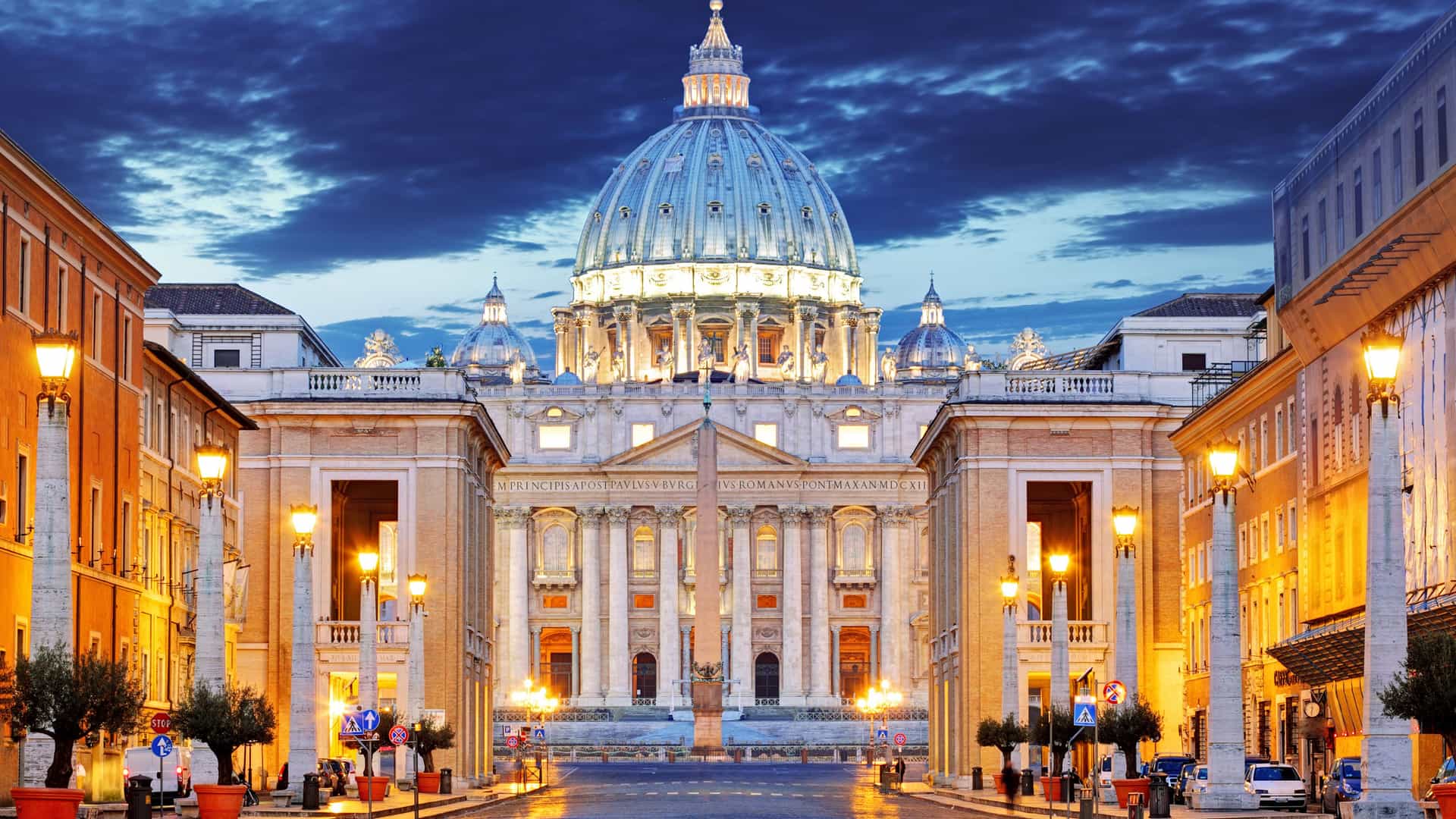 A dramatic night view of St. Peter's Basilica in the Vatican City, a Mediterranean destination. The grand facade and massive dome are beautifully illuminated against the dark blue, cloudy sky. Streetlights line the empty cobblestone street leading up to the basilica, casting a warm yellow glow. The surrounding buildings are also lit up, adding to the majestic and historical atmosphere of this iconic Roman landmark.