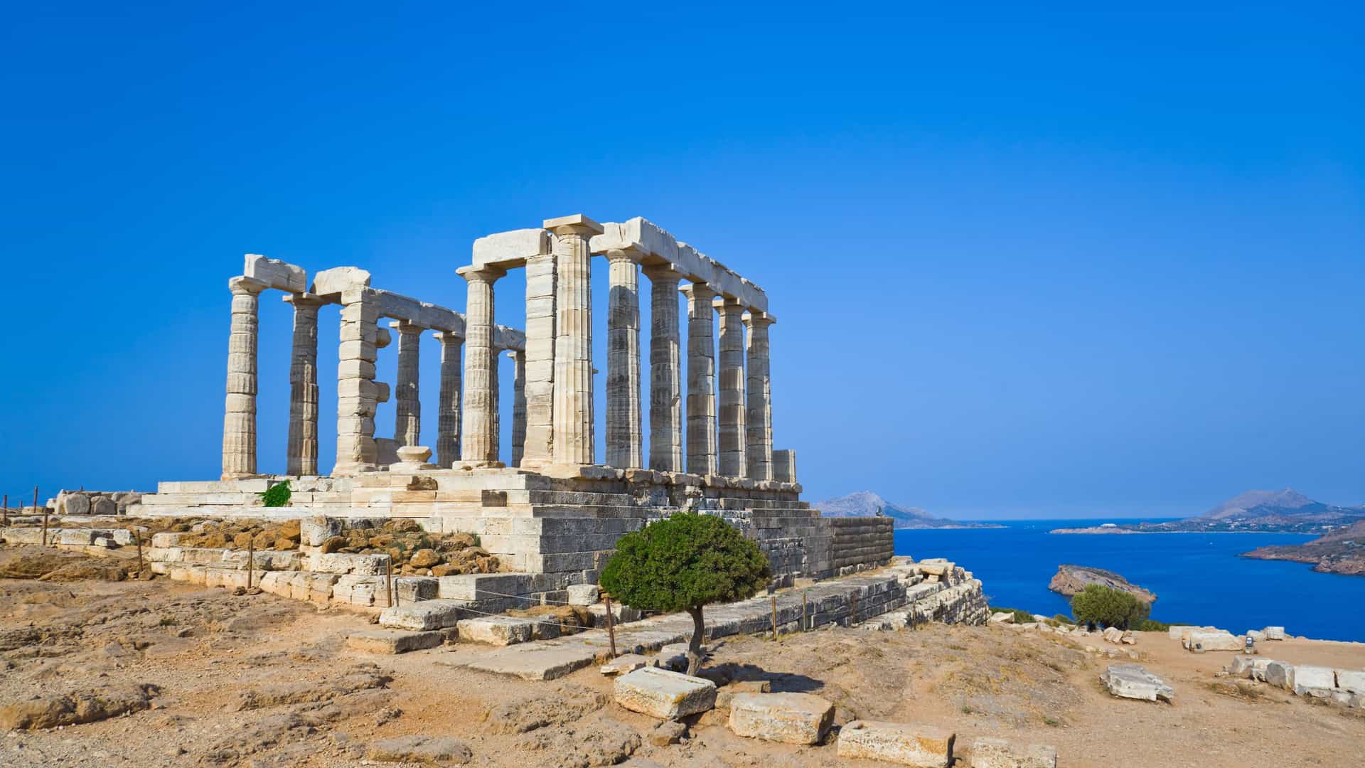 The ancient Temple of Poseidon at Cape Sounion, Greece, overlooking the Aegean Sea. The iconic white marble columns stand against a brilliant, cloudless blue sky and the deep blue water. The temple ruins sit on a rocky headland, with the Mediterranean coastline visible in the distance. The image captures the historical significance and breathtaking seaside location of this Greek landmark.