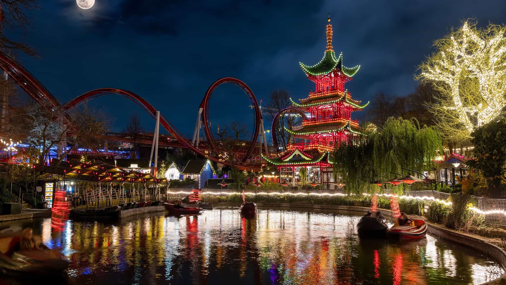 A magical night view of Tivoli Gardens in Copenhagen, Denmark, during the Christmas season. A full moon hangs in the dark sky, illuminating a tranquil pond with small boats. The red-and-green pagoda building is brightly lit with Christmas lights, and a red rollercoaster track is visible in the background. The trees are decorated with twinkling yellow lights, and their reflections shimmer on the water. The image captures the festive and charming atmosphere of this popular Northern European destination at night.