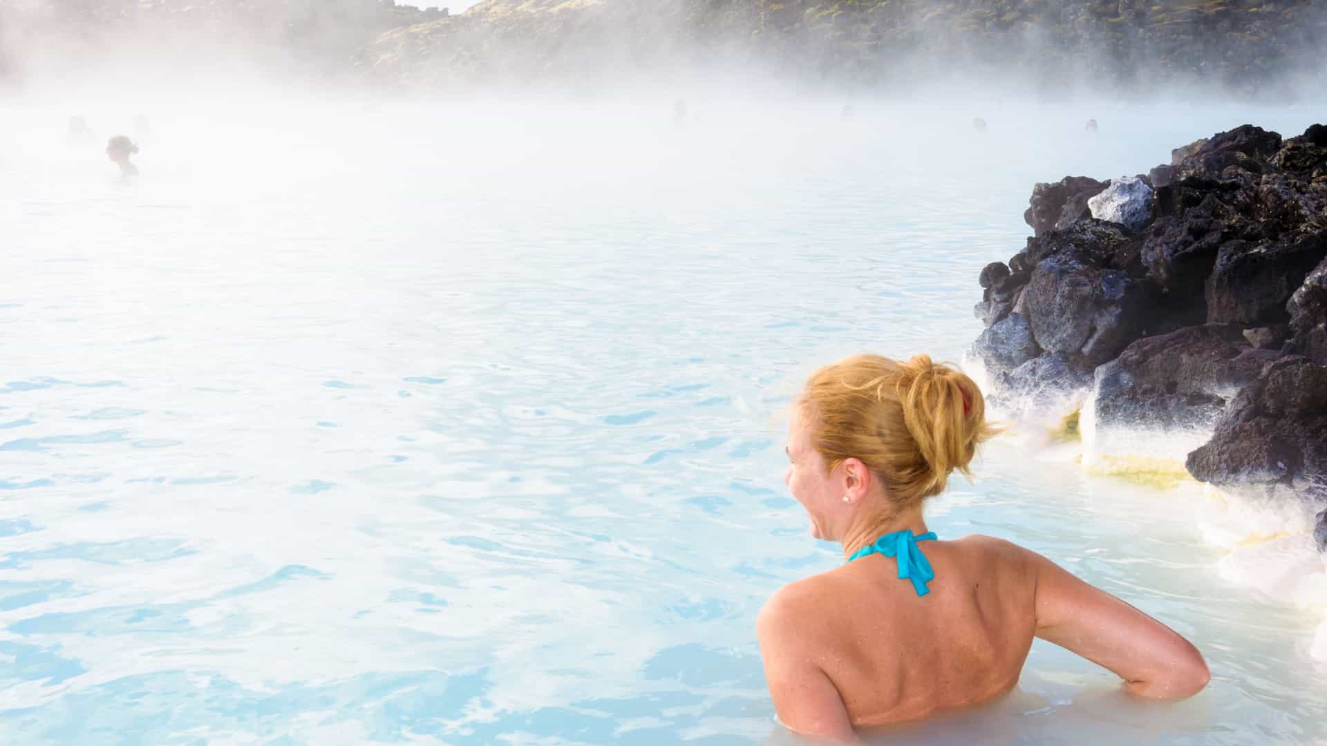 A woman with her back to the camera enjoys the warm, milky-blue waters of the Blue Lagoon geothermal spa in Iceland. Her blonde hair is pulled back, and she wears a blue bikini top. Steam rises from the water, creating a misty atmosphere. The lagoon is bordered by dark, volcanic rocks, and other indistinct figures are visible in the foggy background. The image captures the serene and unique spa experience in this popular Northern European destination.