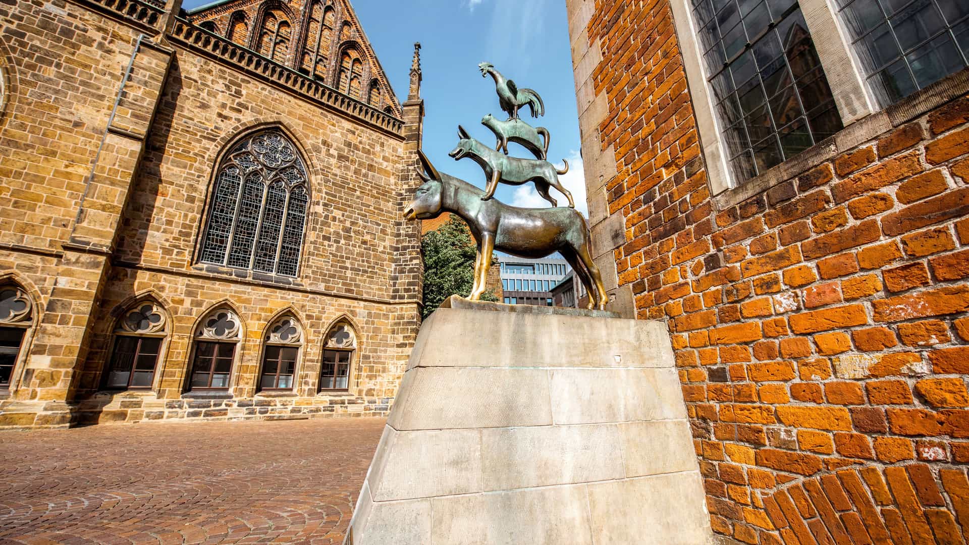 The famous 'Bremen Town Musicians' statue in Bremen, Germany. The bronze sculpture, based on the Brothers Grimm fairy tale, features a donkey, a dog, a cat, and a rooster stacked on top of each other. The statue sits on a stone pedestal in a cobblestone square. In the background, a large, ornate brick building, likely Bremen Cathedral, is partially visible. The clear blue sky and old European architecture highlight this Northern European fairytale landmark.