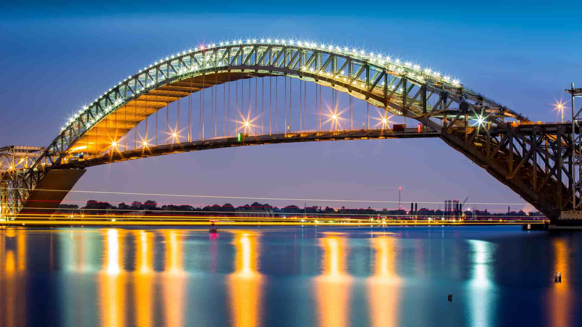 A night-time view of the iconic Bayonne Bridge in New Jersey, brightly lit against a dark sky, reflecting its lights on the still water of the Kill Van Kull waterway.