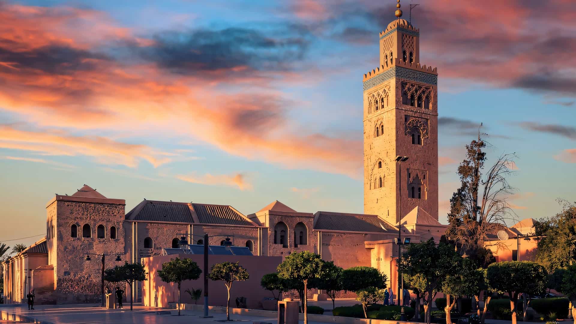 "A wide-angle sunset view of the Koutoubia Mosque in Marrakech, Morocco, with its prominent minaret silhouetted against a vibrant orange and blue sky. "