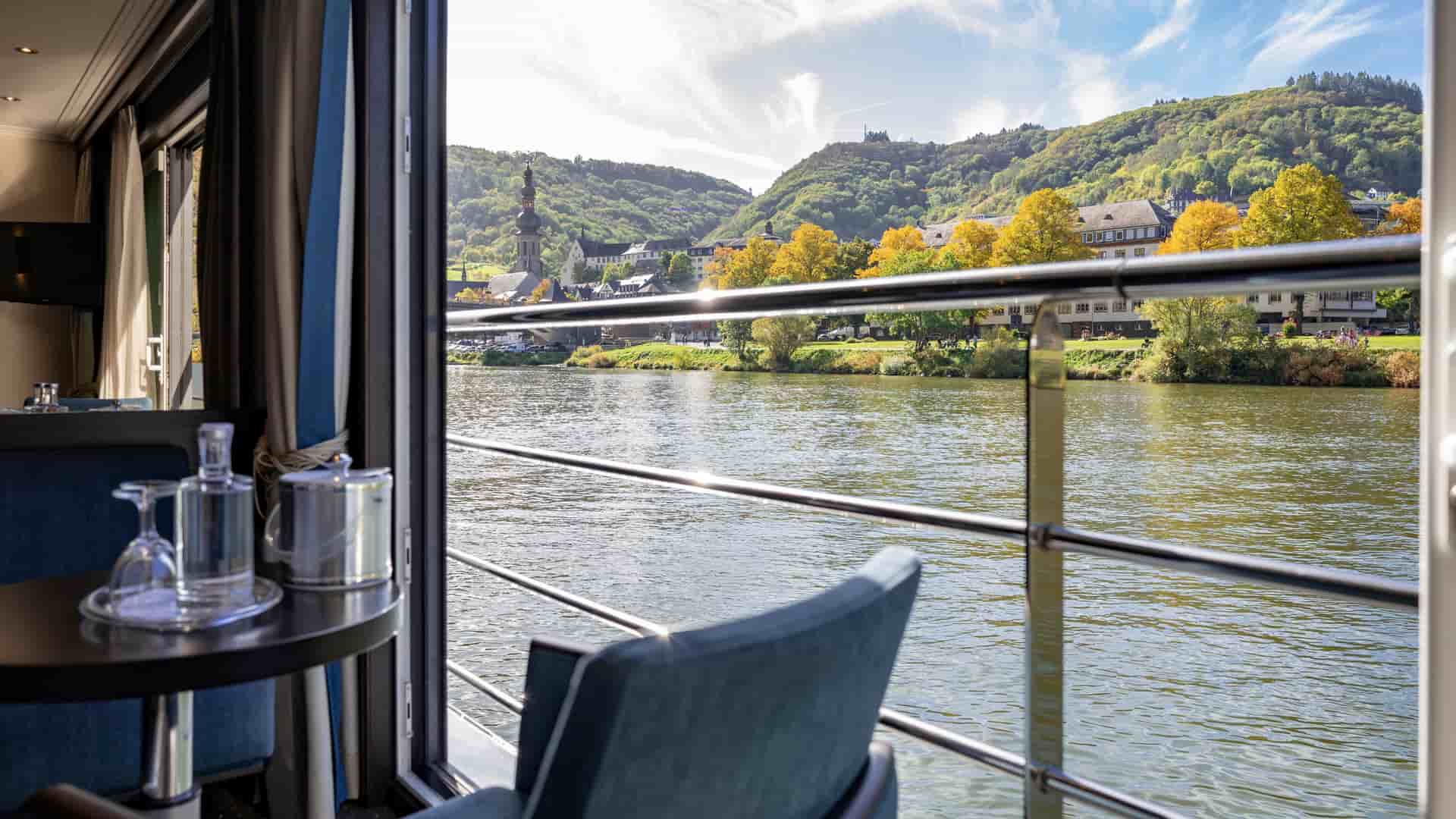 Open-air Panorama Suite aboard an Avalon Waterways ship featuring a wide sliding glass door framing a scenic river, a balcony railing, and a European hillside village with a church steeple.