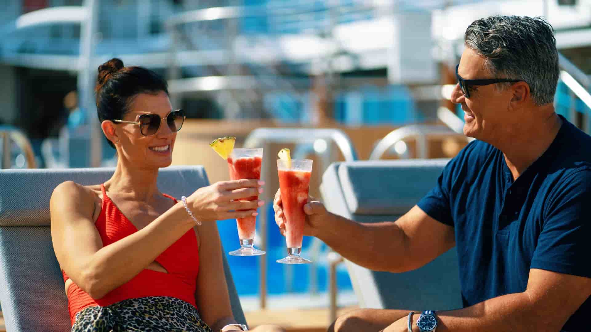 A man and woman on the Azamara Quest ship are sitting in lounge chairs on an outdoor deck, toasting each other with red and yellow cocktails with pineapple slices.