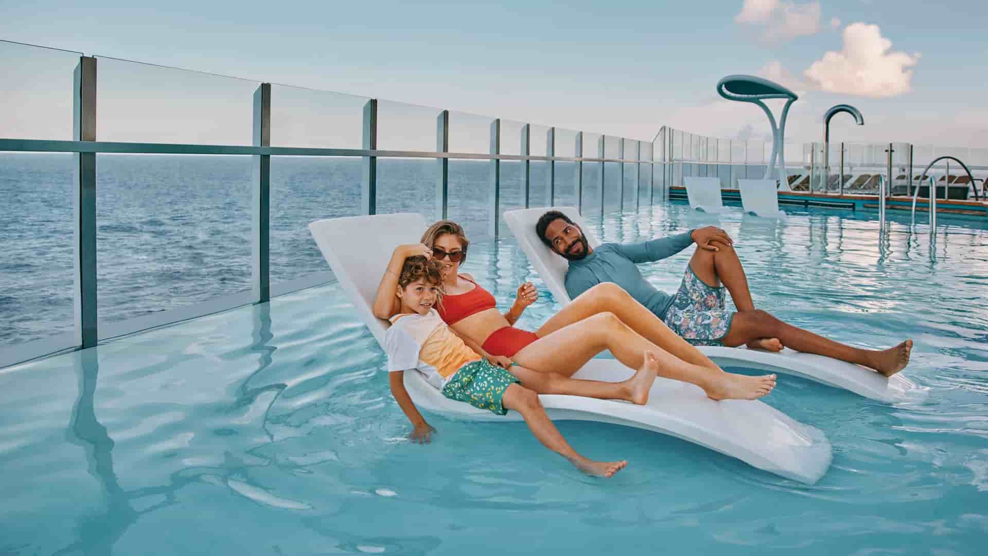 A family of three is lounging in a shallow infinity pool on the Carnival Mardi Gras ship, which looks out over the ocean. The mother and young son are on one white lounge chair, and the father is on another.