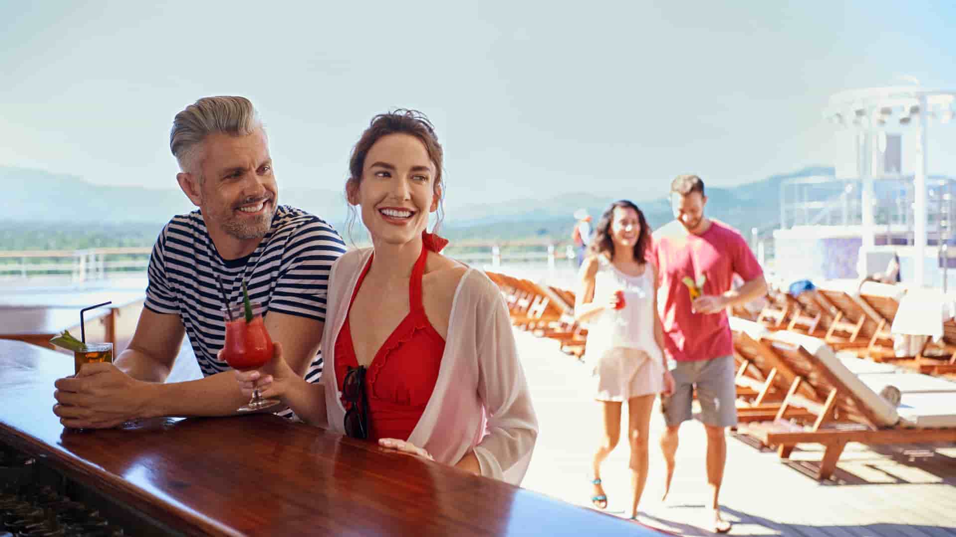 A smiling couple enjoying cocktails at a outdoor bar onboard a Cunard cruise ship during a bright, sunny day.