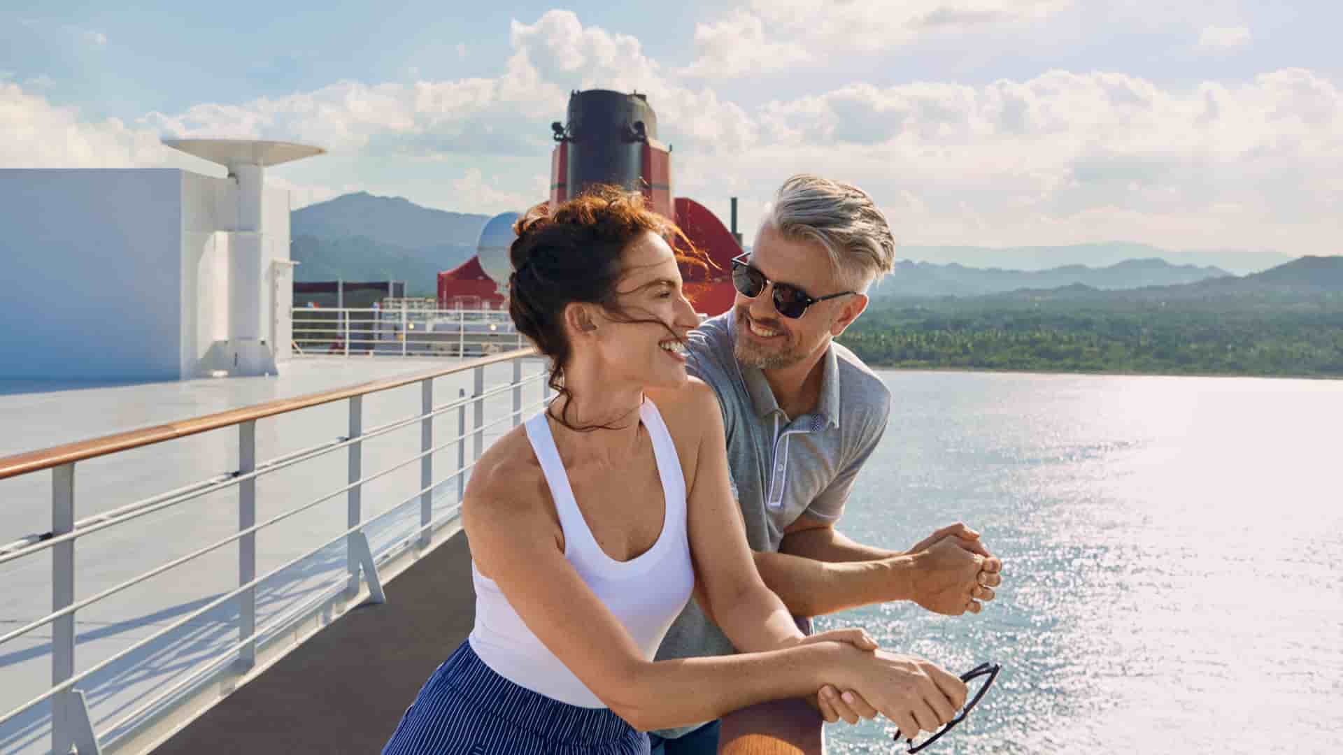 A couple on the deck of the Cunard Queen Mary 2 ship, with a man leaning on the rail looking at the laughing woman, with scenic mountains and the ocean in the background.
