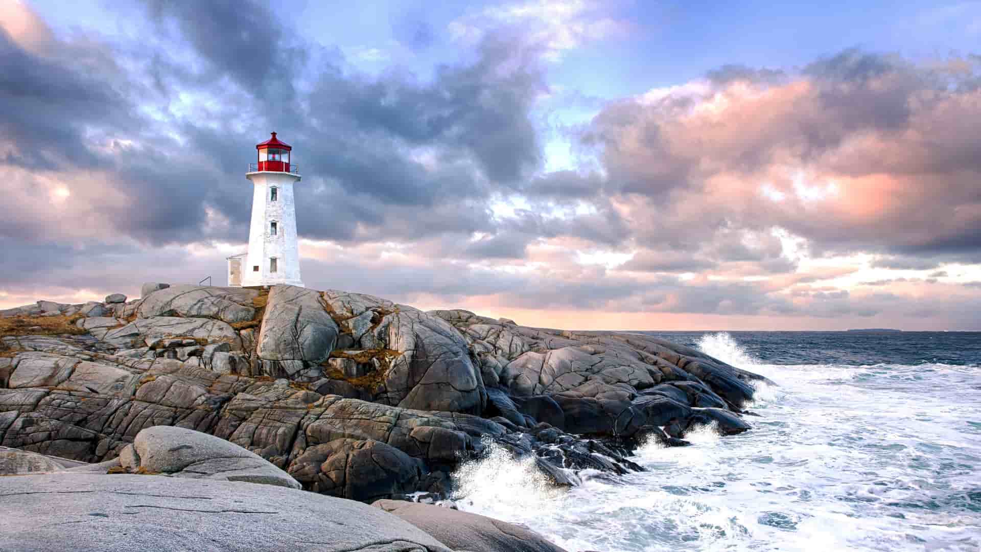 Peggy's Cove Lighthouse stands on rocky granite shore with waves crashing against rocks under a dramatic cloudy sky at sunset.