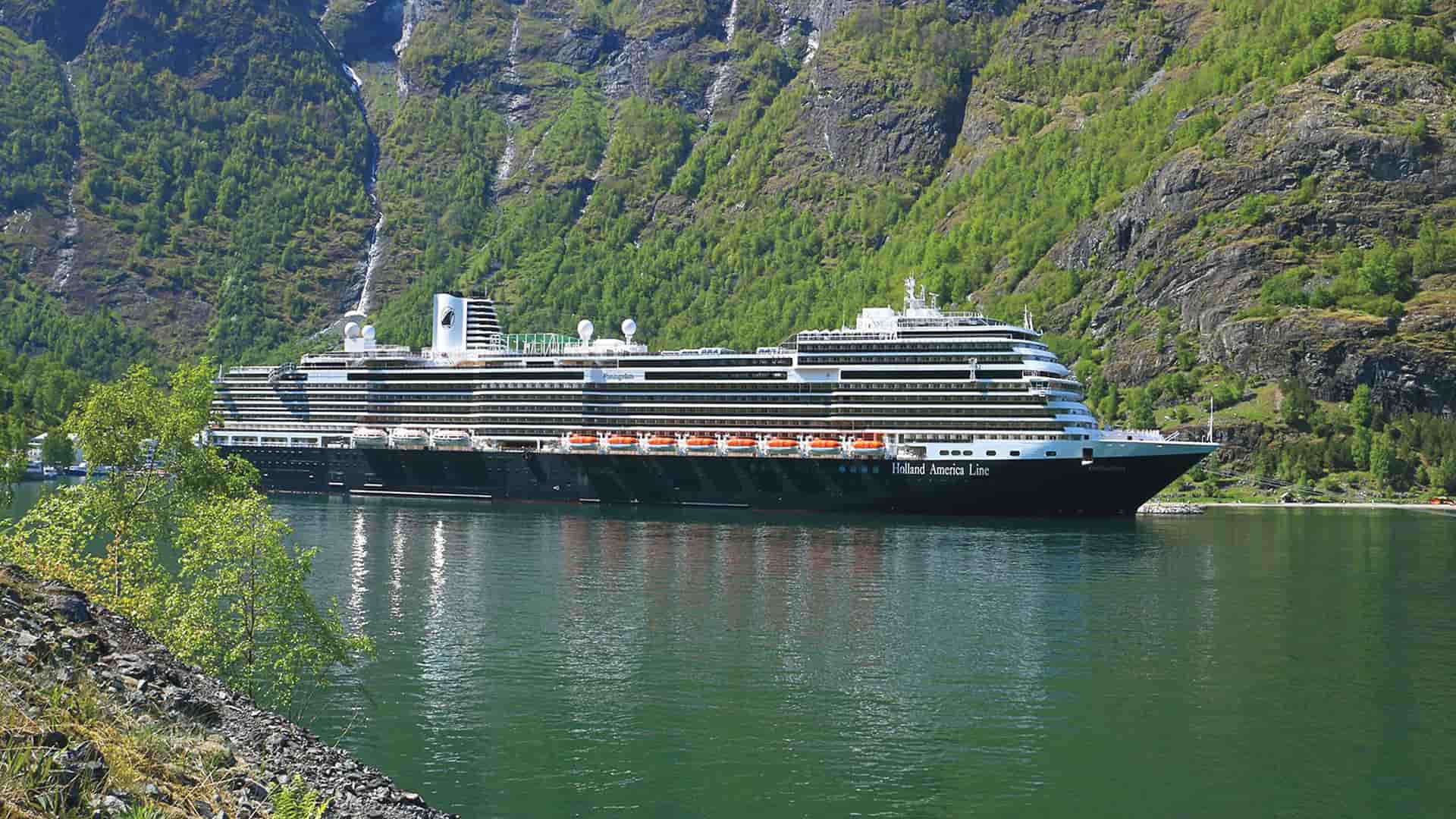 Holland America's Koningsdam at sea by a lush, green mountainside.