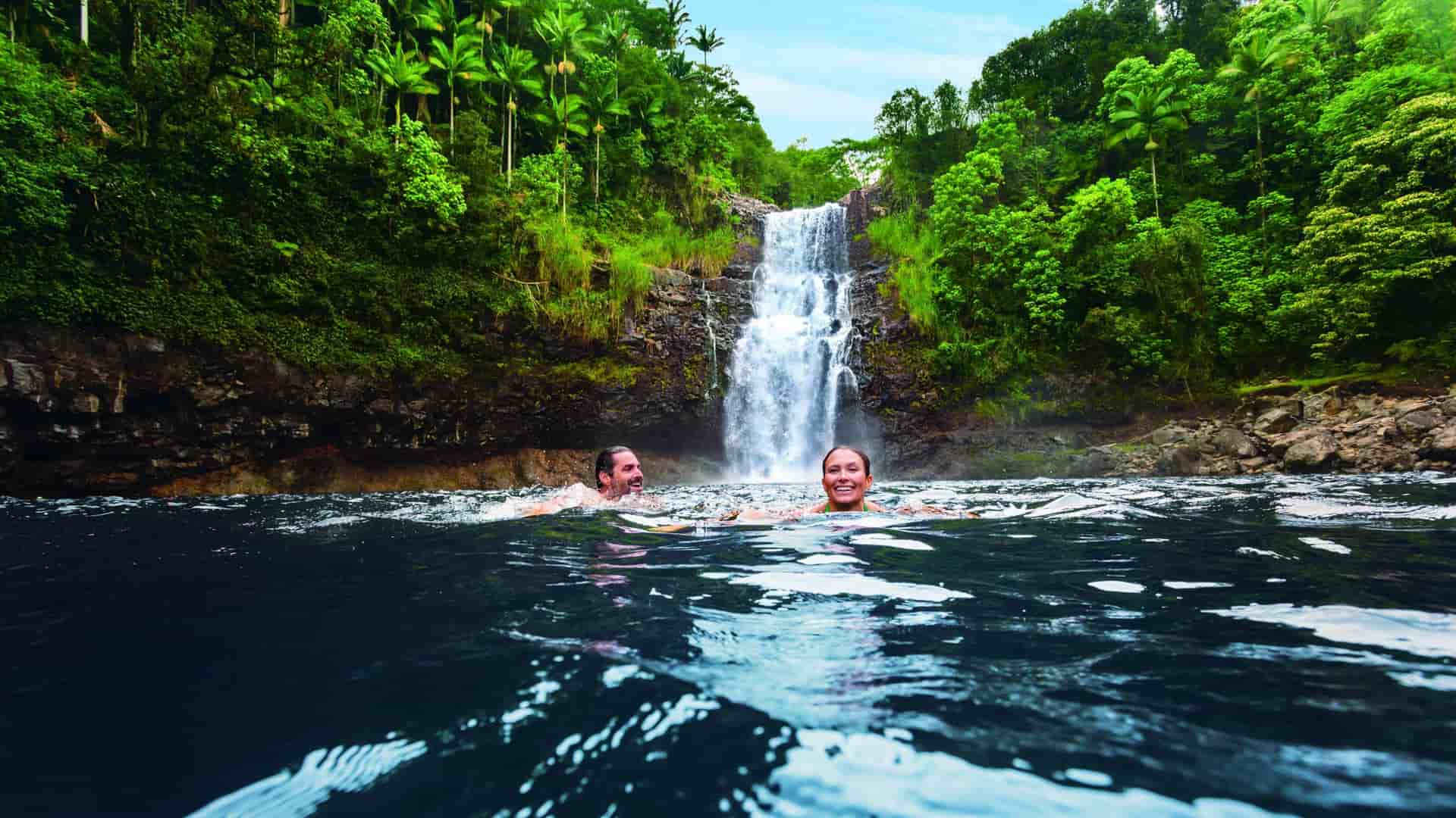 A smiling couple, a man and a woman, swims in a pool of water below a tropical Hawaiian waterfall. The surrounding area is lush with green trees and foliage.