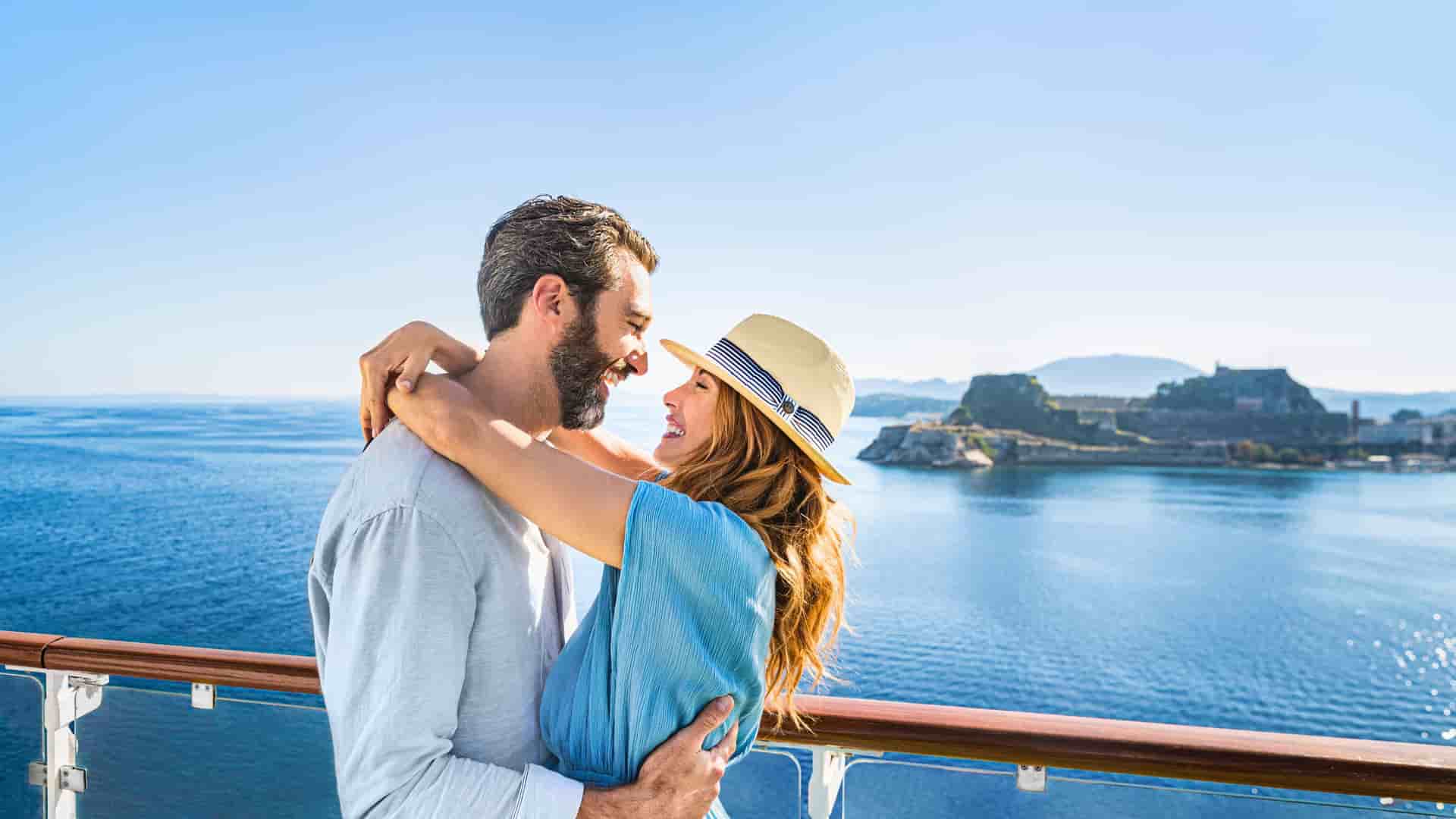 A happy couple embraces on the deck of a Norwegian Cruise Line ship, with a panoramic view of the coastline and Old Fortress of Corfu, Greece, in the background.