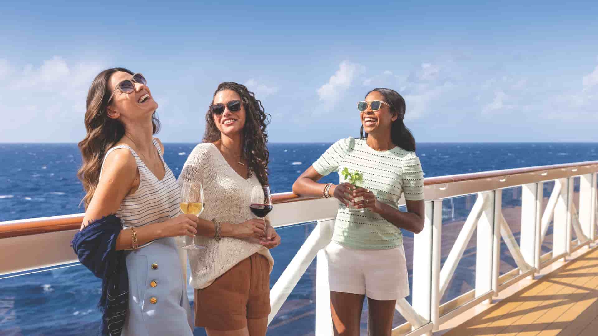 3 women laughing together while leading on the deck railing on Norwegian Encore during a bright, sunny day at sea.