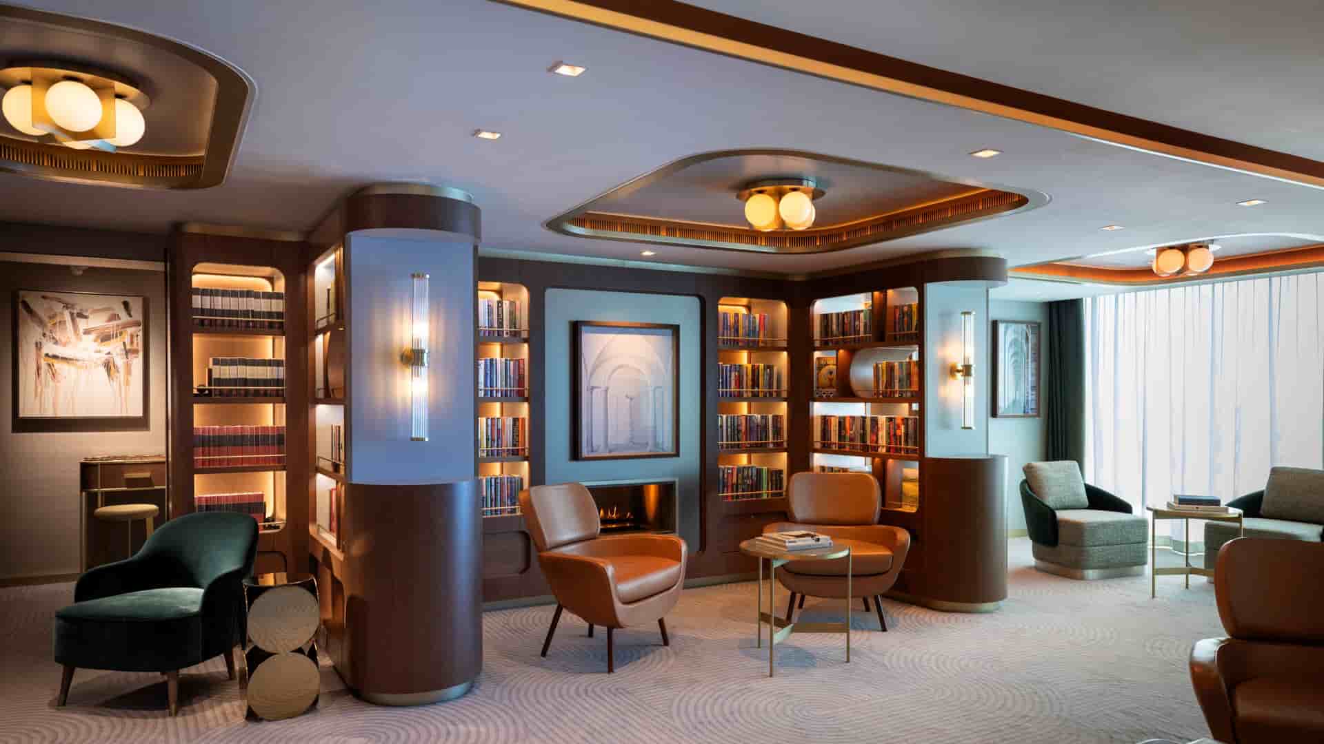 An elegantly designed ship library featuring built-in dark wood bookshelves, comfortable orange leather and green velvet armchairs, warm recessed lighting, and a patterned carpet.