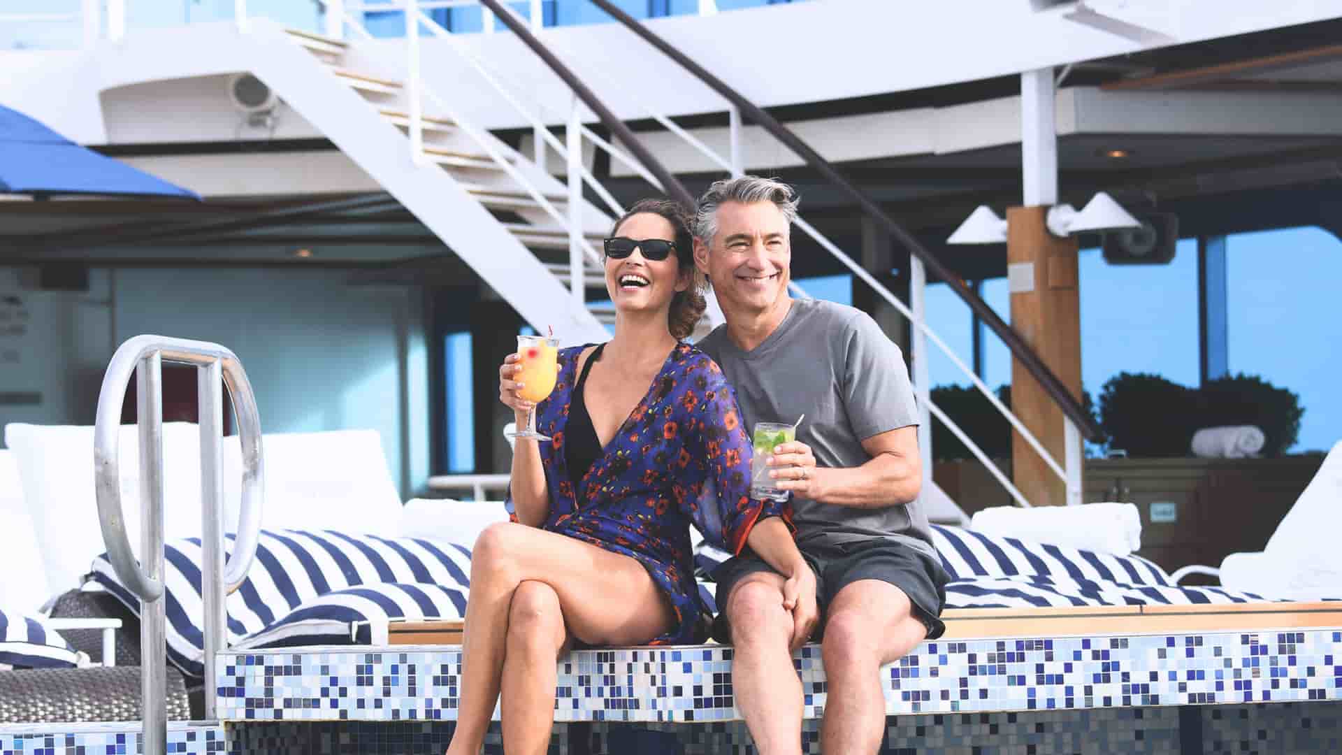 A happy couple relaxes with cocktails on the mosaic-tiled pool deck of the Oceania Riviera cruise ship, showcasing a leisurely onboard experience.