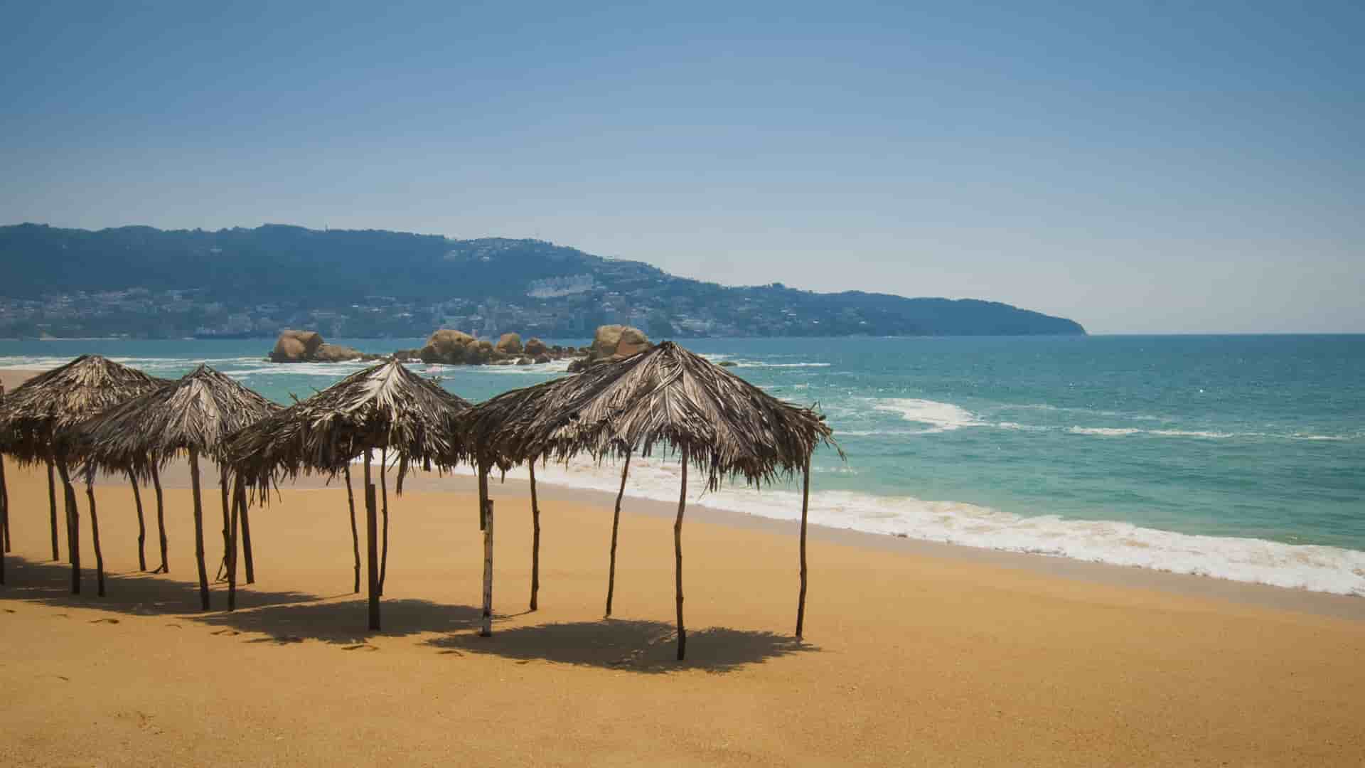 A beautiful beach in Acapulco, Mexico, with thatched-roof umbrellas on the golden sand, facing the turquoise ocean under a clear blue sky.