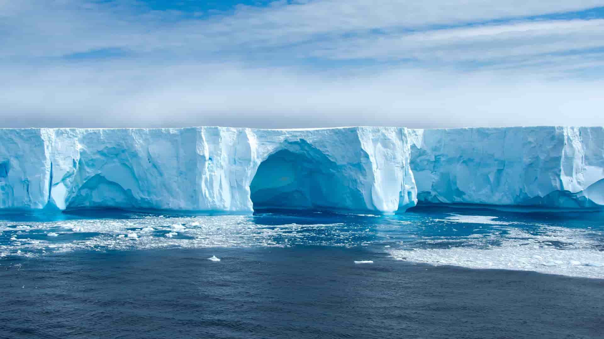 A majestic view of a massive, glowing blue iceberg with natural archways and an icy shoreline in Admiralty Bay, Antarctica, under a dramatic cloudy sky.