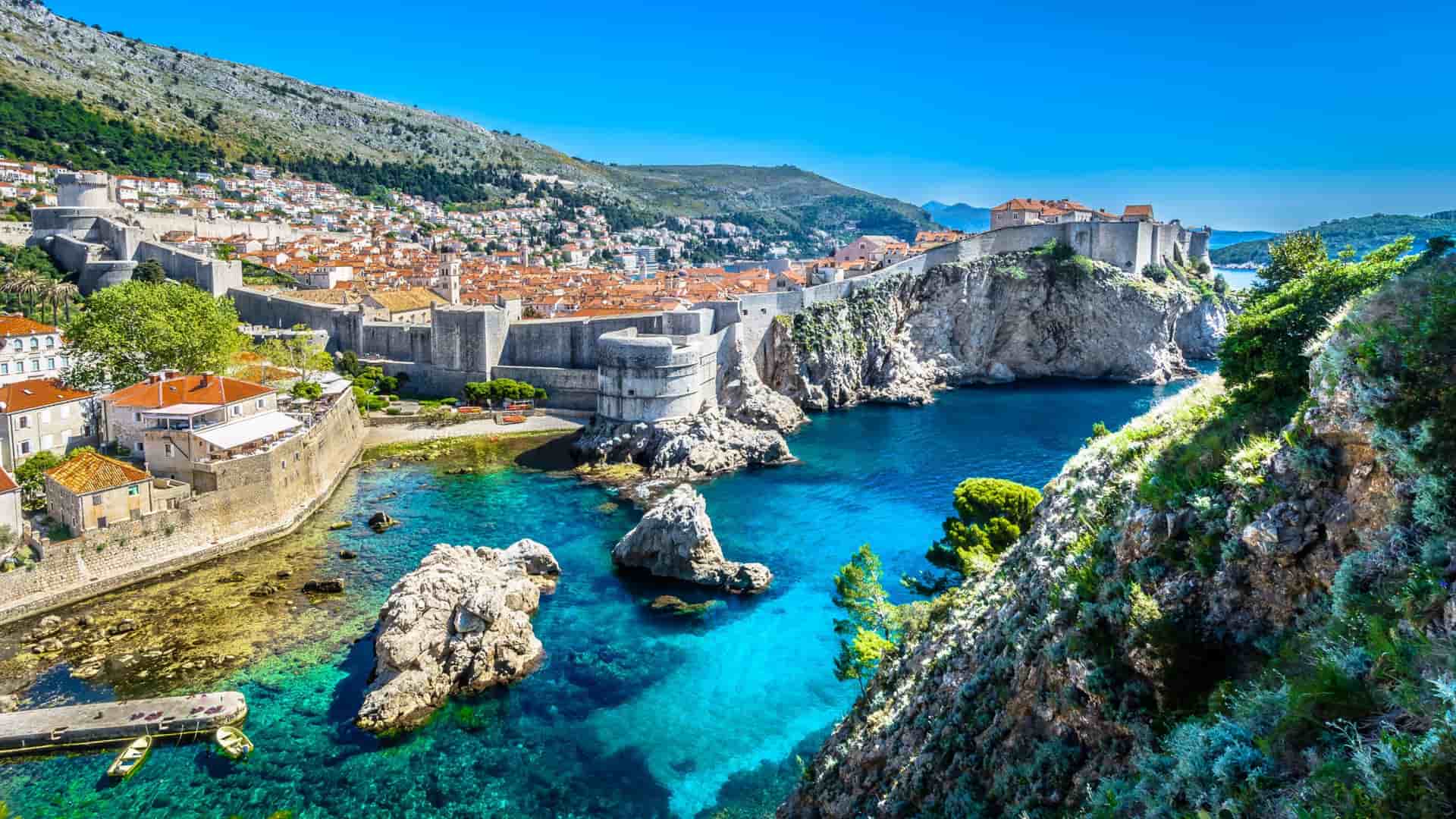 A scenic view of Dubrovnik's Old Town, with its medieval city walls and terracotta roofs, perched on cliffs overlooking the clear turquoise waters of the Adriatic Sea.