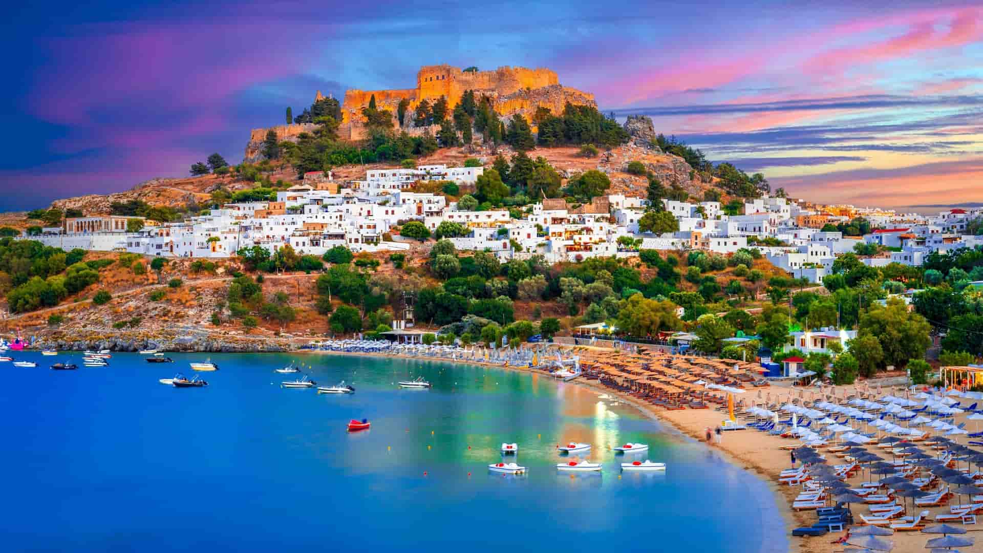 A panoramic sunset view of Lindos on the Greek island of Rhodes, with its iconic Acropolis and whitewashed houses overlooking a bay of the Aegean Sea.
