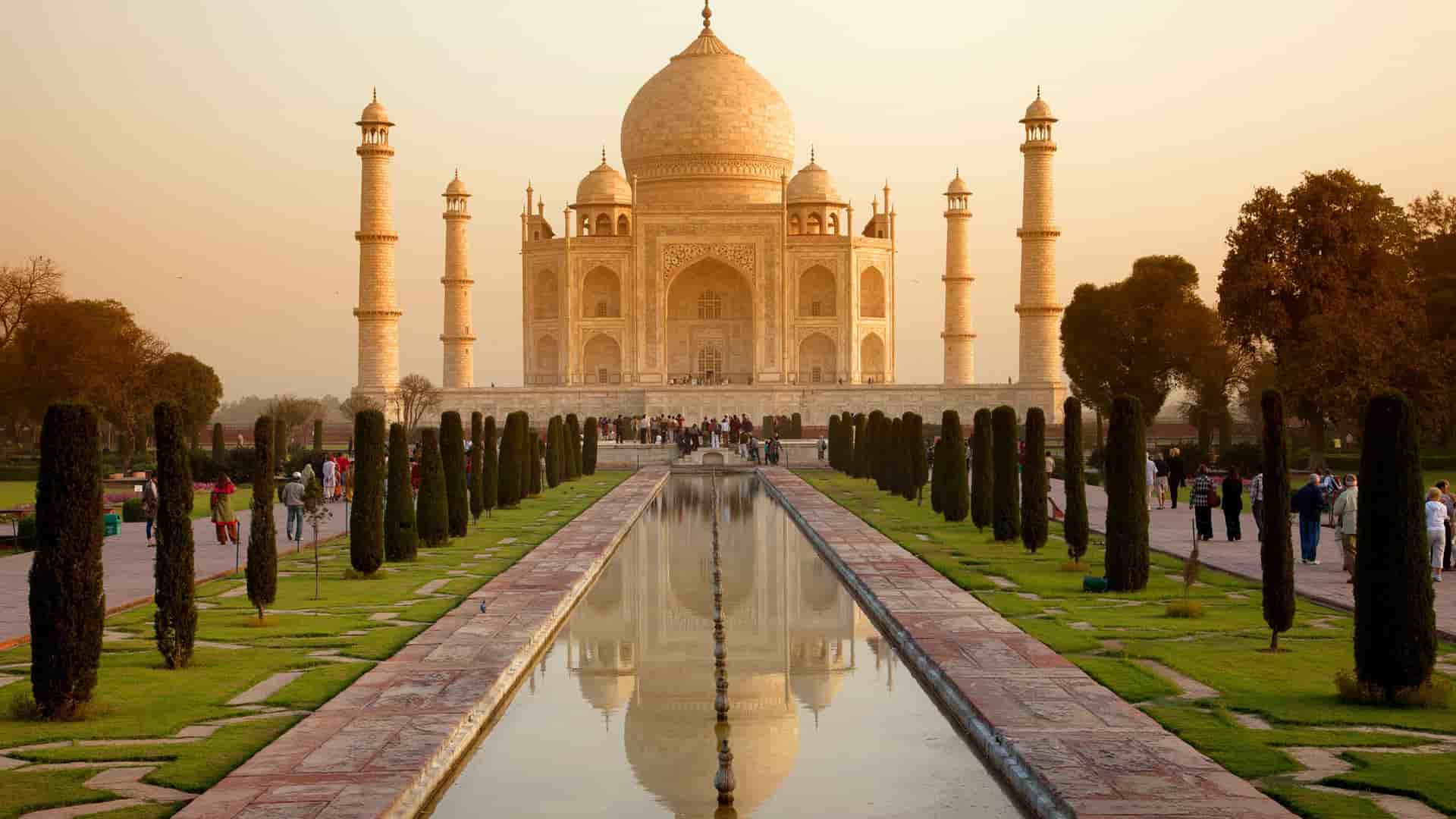 A majestic view of the Taj Mahal in Agra, India, with its white marble dome and minarets reflected in the long, symmetrical pool at sunset.