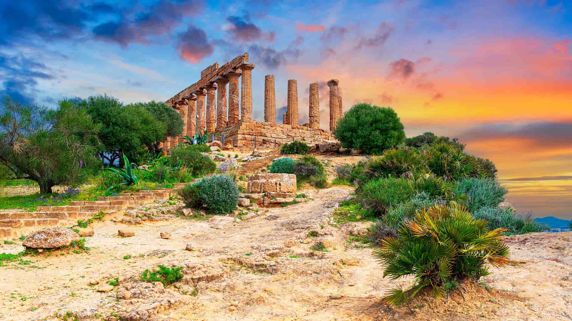 The ancient Greek temple ruins in Agrigento, Italy, stand on a rocky hillside under a dramatic, colorful sunset sky. The well-preserved ruins, consisting of a row of tall, fluted stone columns, are surrounded by green shrubs and a few small trees.