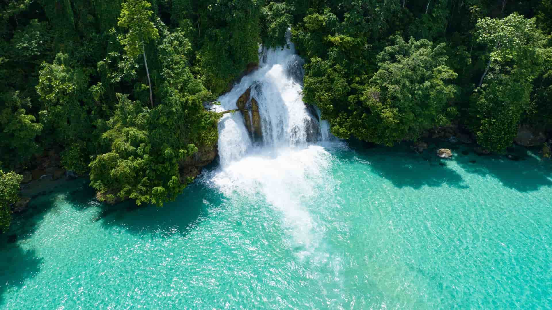 An aerial view of Air Terjun Kiti, a powerful waterfall cascading into a stunningly clear turquoise pool, surrounded by dense tropical rainforest.