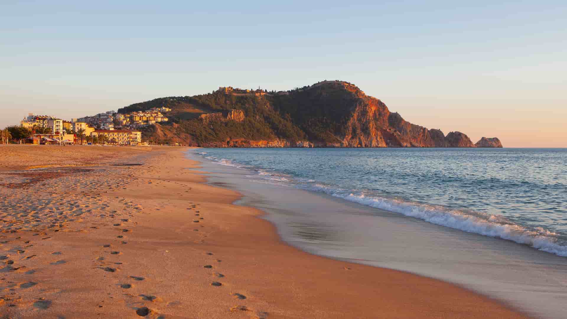 A scenic sunset view of Cleopatra Beach in Alanya, Turkey, with a long sandy shore and gentle waves, a rugged cliff face, and the Alanya Castle on top.