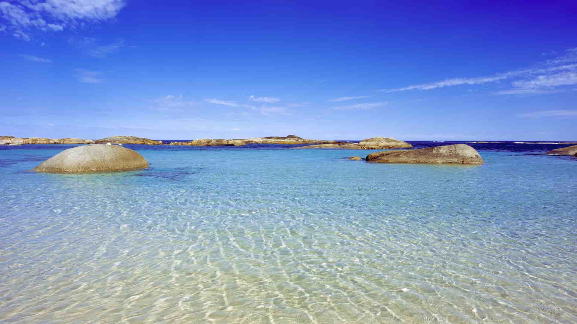 A scenic view of Elephant Rocks beach in Albany, Western Australia, with massive granite boulders dotting the shallow, crystal-clear turquoise water under a bright blue sky.