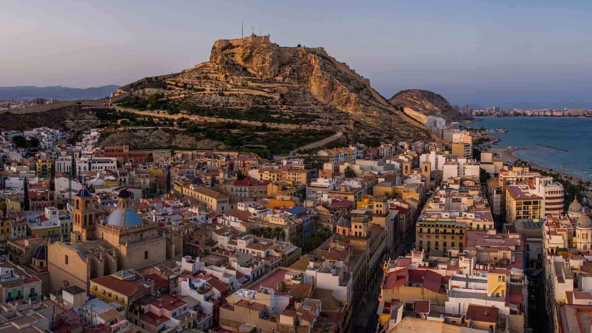 A stunning panoramic view of Alicante, Spain, at sunset, with the historic city nestled at the foot of Mount Benacantil and the Santa Bárbara Castle, with the Mediterranean Sea in the distance.