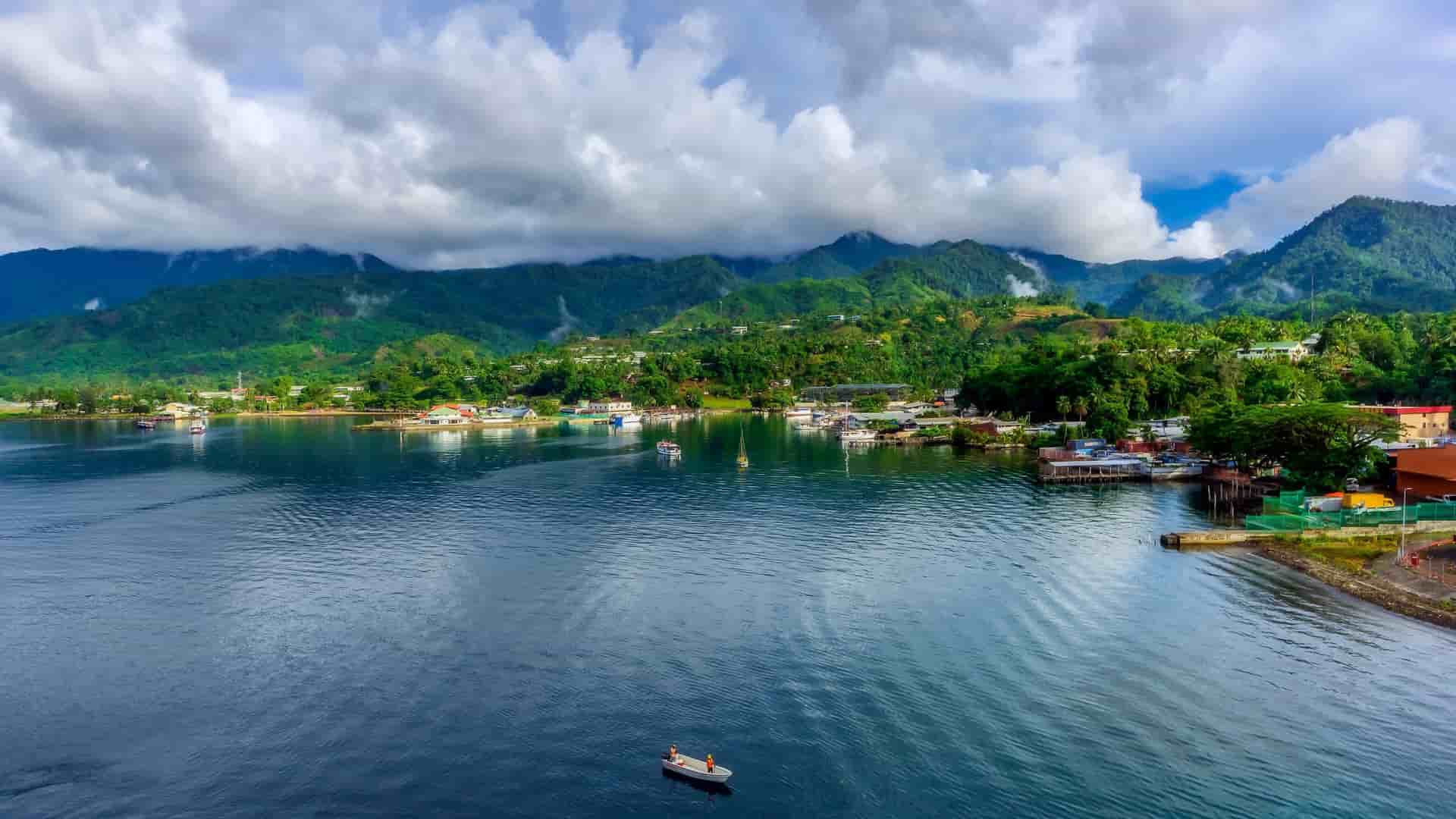 A scenic view of Alotau, Papua New Guinea, with a vibrant harbor and a small town nestled against a backdrop of lush green mountains under a cloudy sky.