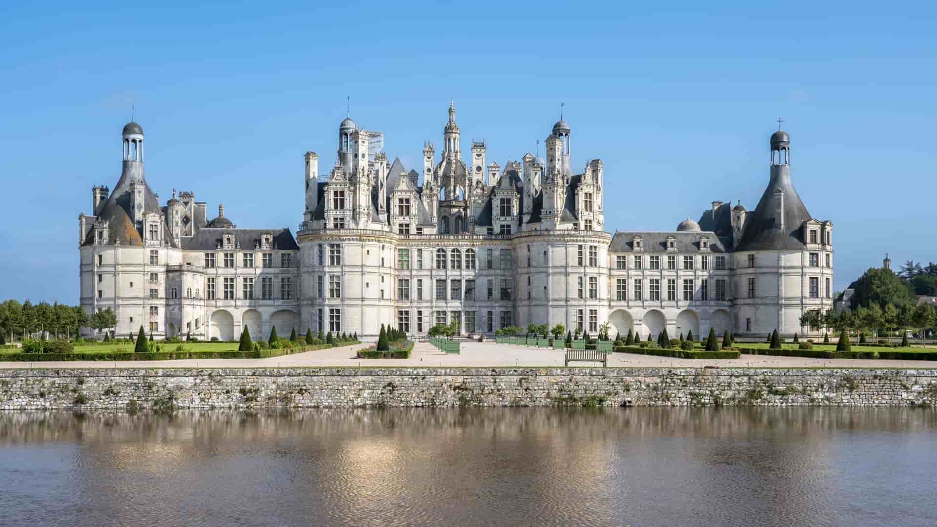 A magnificent wide-shot of the historic Château de Chambord, a Renaissance castle with ornate turrets, reflected in the still waters of the Loire River in Amboise, France.