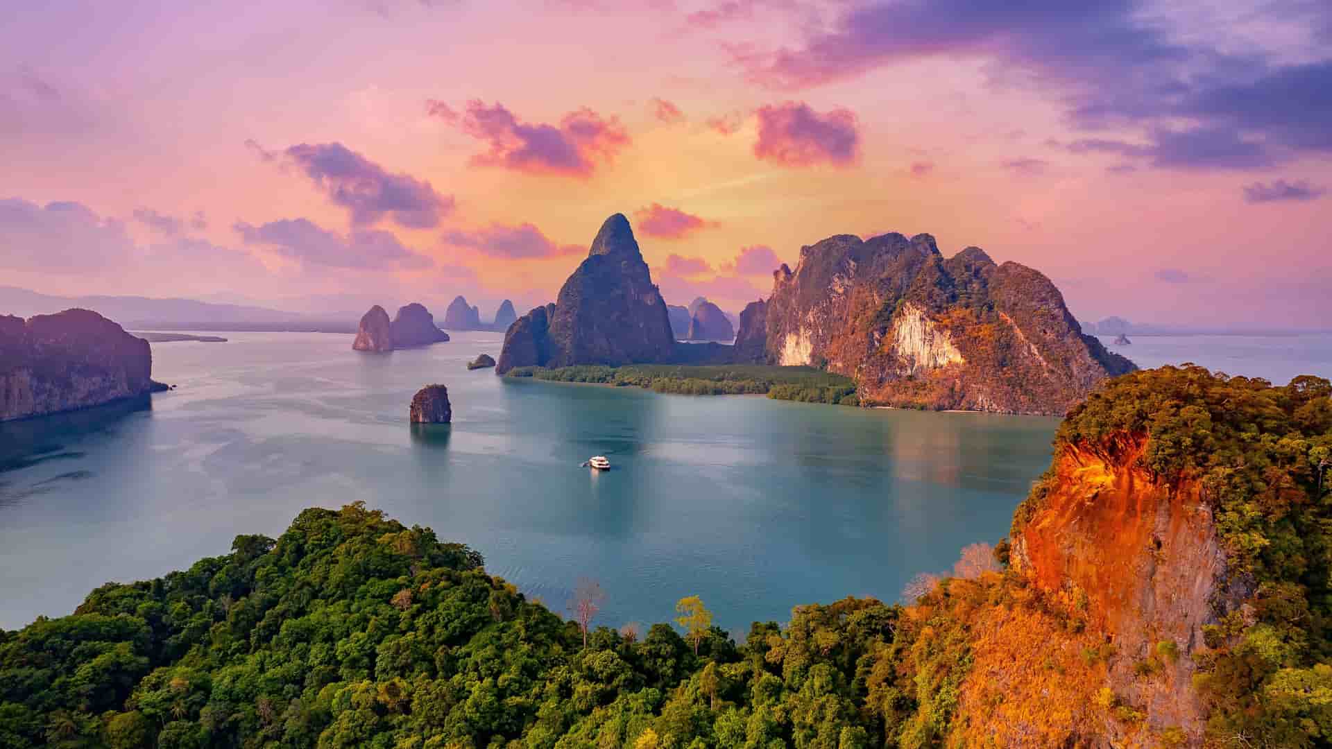 A wide-angle sunset view of the Andaman Sea's iconic limestone cliffs and rock formations rising from the serene waters, with a boat sailing in the foreground.