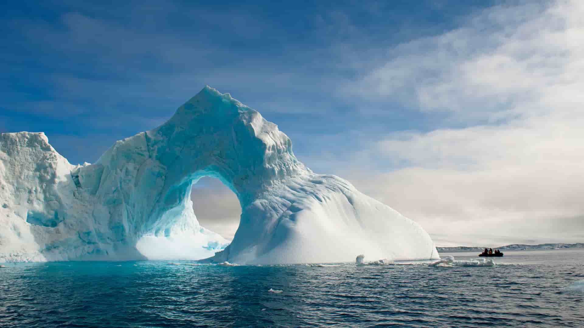 A zodiac boat carrying passengers cruises near a massive, naturally formed archway in an iceberg, showcasing the stunning scenery of the Antarctic Sound.