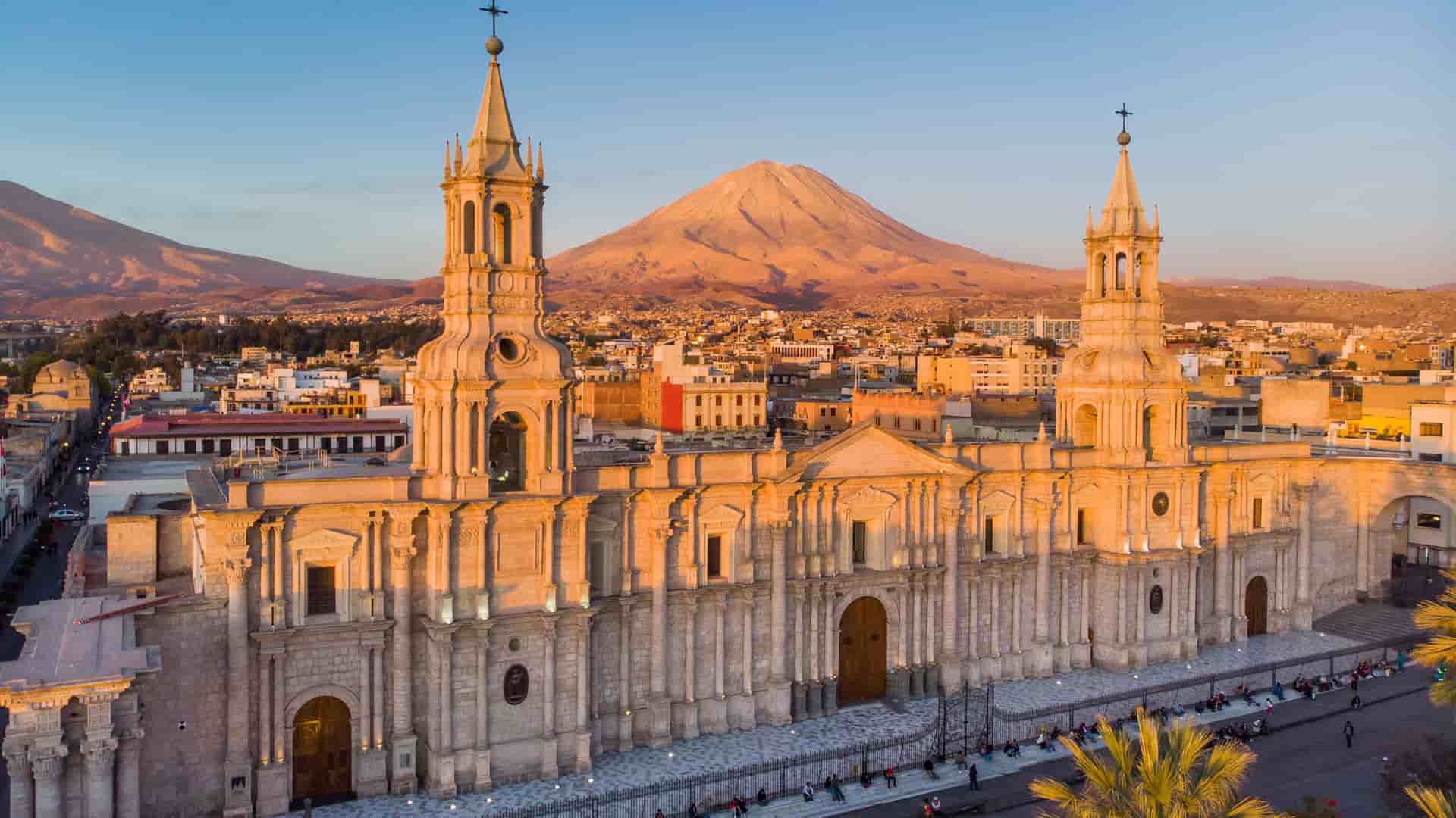 The monumental white facade of the Basilica Cathedral of Arequipa in Peru is bathed in the golden light of sunset. In the background, the symmetrical cone of the majestic Misti volcano rises above the city skyline under a clear, warm-toned sky.