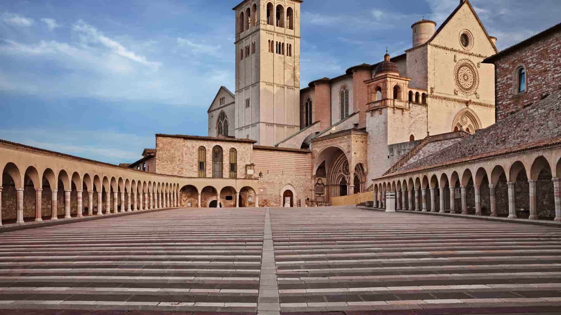 The Basilica of Saint Francis of Assisi in Italy, a large historic building with a prominent bell tower, rises above a vast paved courtyard with a striped geometric pattern. The courtyard is lined on both sides by arched colonnades, leading the eye toward the basilica under a partly cloudy sky.