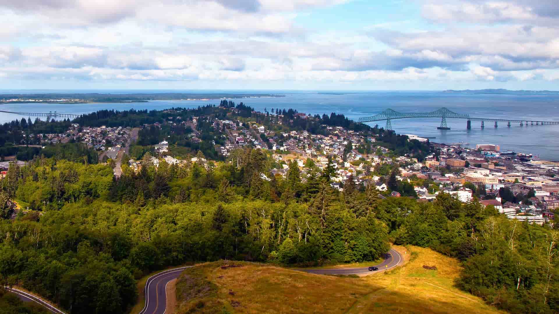 An aerial view of Astoria, Oregon, showcasing a winding road, a lush green hill, the city's residential area, and the Astoria-Megler Bridge crossing the Columbia River.