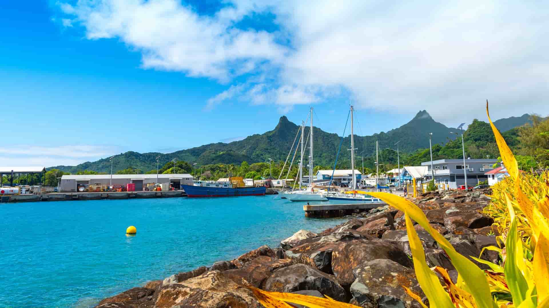 A picturesque view of Avatiu Harbour in Rarotonga, Cook Islands, with boats docked in the turquoise water, a rocky shoreline, and lush green mountains in the background.