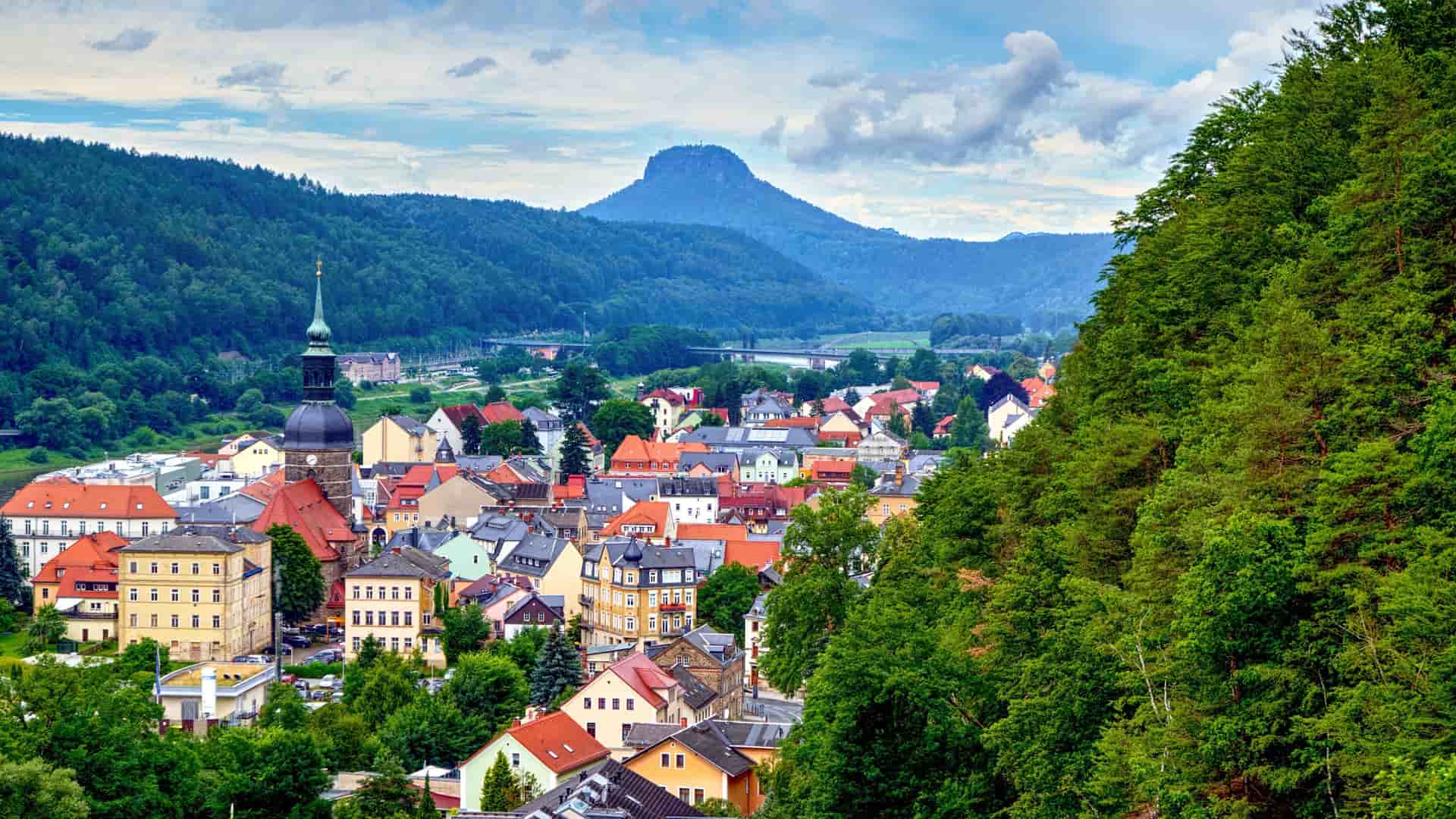 A picturesque view of the village of Bad Schandau in Germany, with colorful buildings and a church steeple in the foreground and a prominent mountain peak in the background.