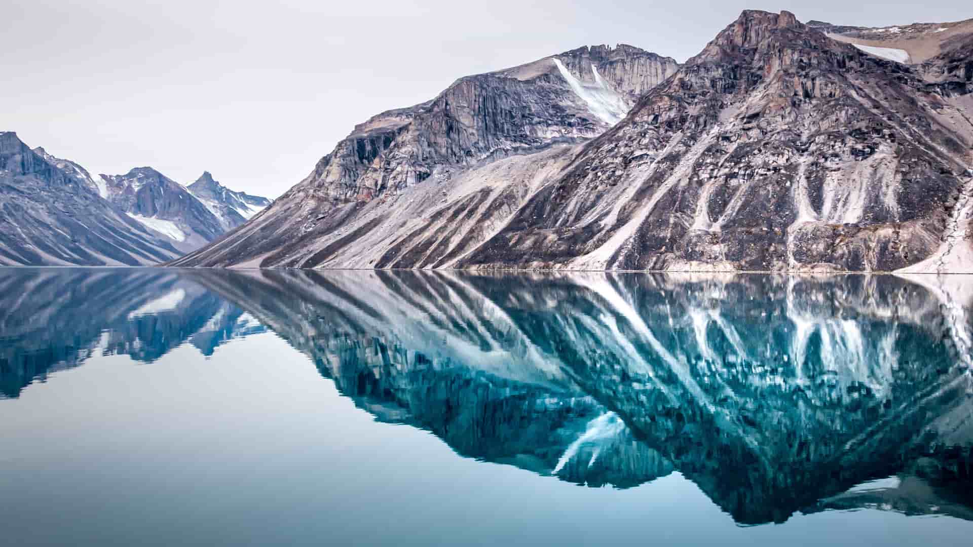 A breathtaking view of dramatic, rugged mountains in Baffin Island, Canada, with snow and striated rock on their slopes. The mountains are perfectly reflected in the still, glassy surface of a large body of water, creating a serene and symmetrical natural landscape under a pale sky.
