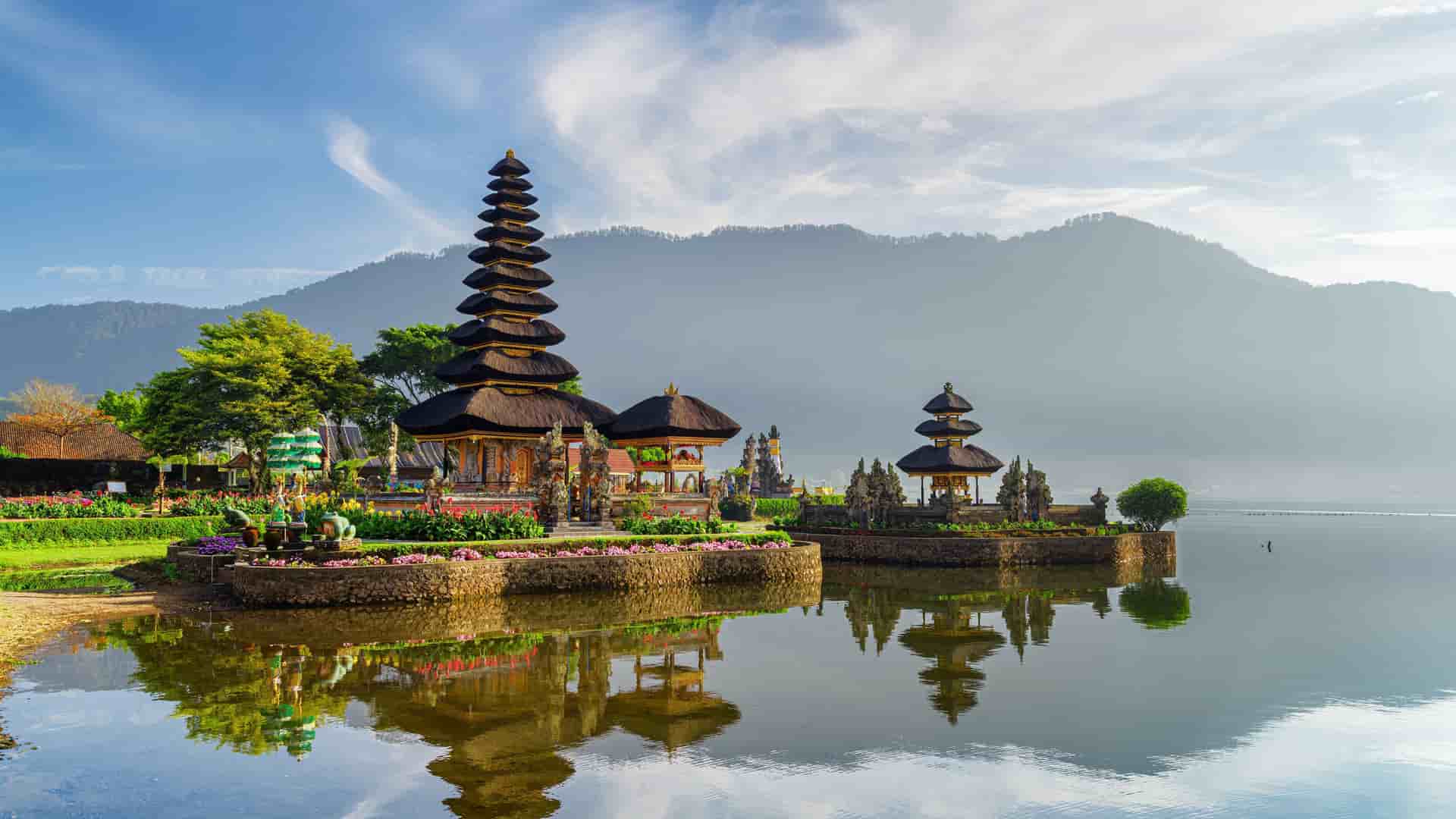 A scenic view of the Ulun Danu Beratan Temple, a Hindu temple on the shores of Lake Beratan in Bali, with tiered pagodas reflecting in the calm water and mountains in the background.