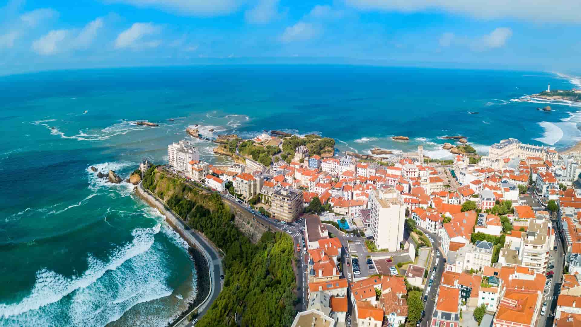 An aerial panoramic view of the Bay of Biscay coastline, showing the red-roofed buildings of Biarritz, France, along the ocean with waves crashing onto a rocky beach.