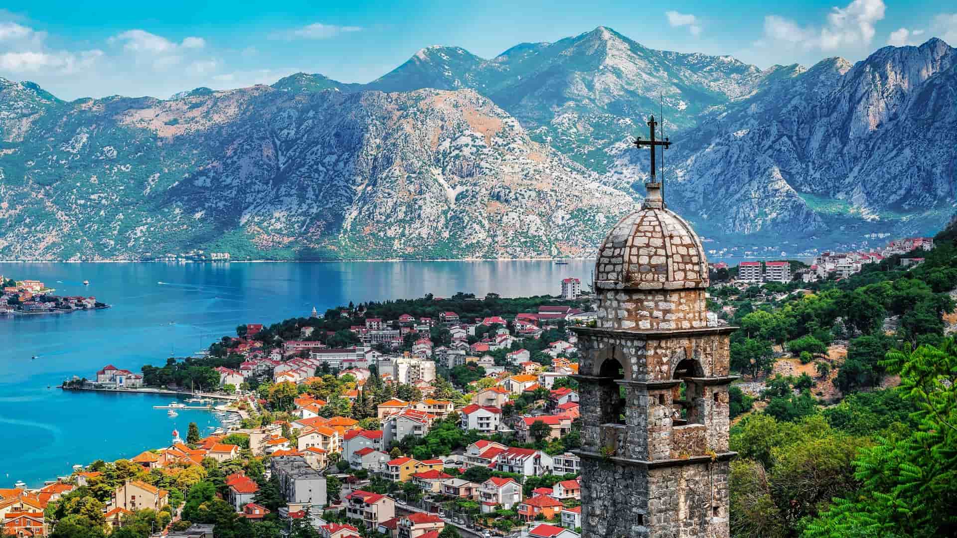 A panoramic view of the Bay of Kotor with a medieval church steeple in the foreground, overlooking the scenic coastal town nestled among towering mountains.